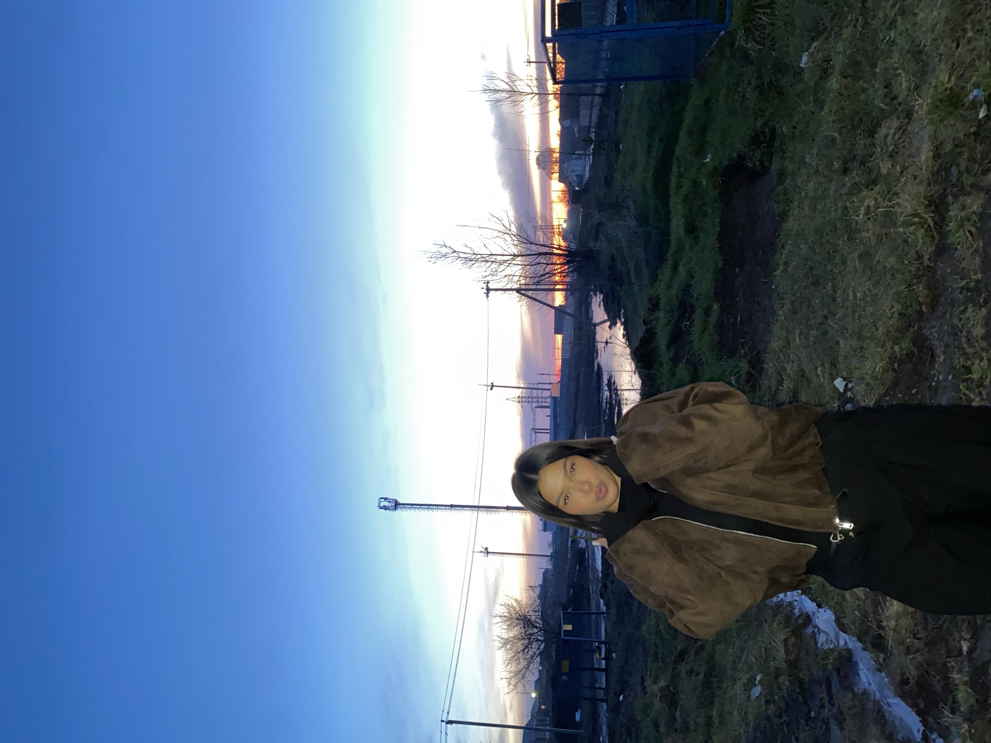 A woman in a brown jacket and black pants is photographed in Chaikovsky, Russia, at dusk. She is positioned in front of a landscape that includes power lines, bare trees, and a body of water reflecting the fading light. In the background, there is a fence and some vegetation. The sky transitions from blue to orange, indicating a sunset or sunrise.