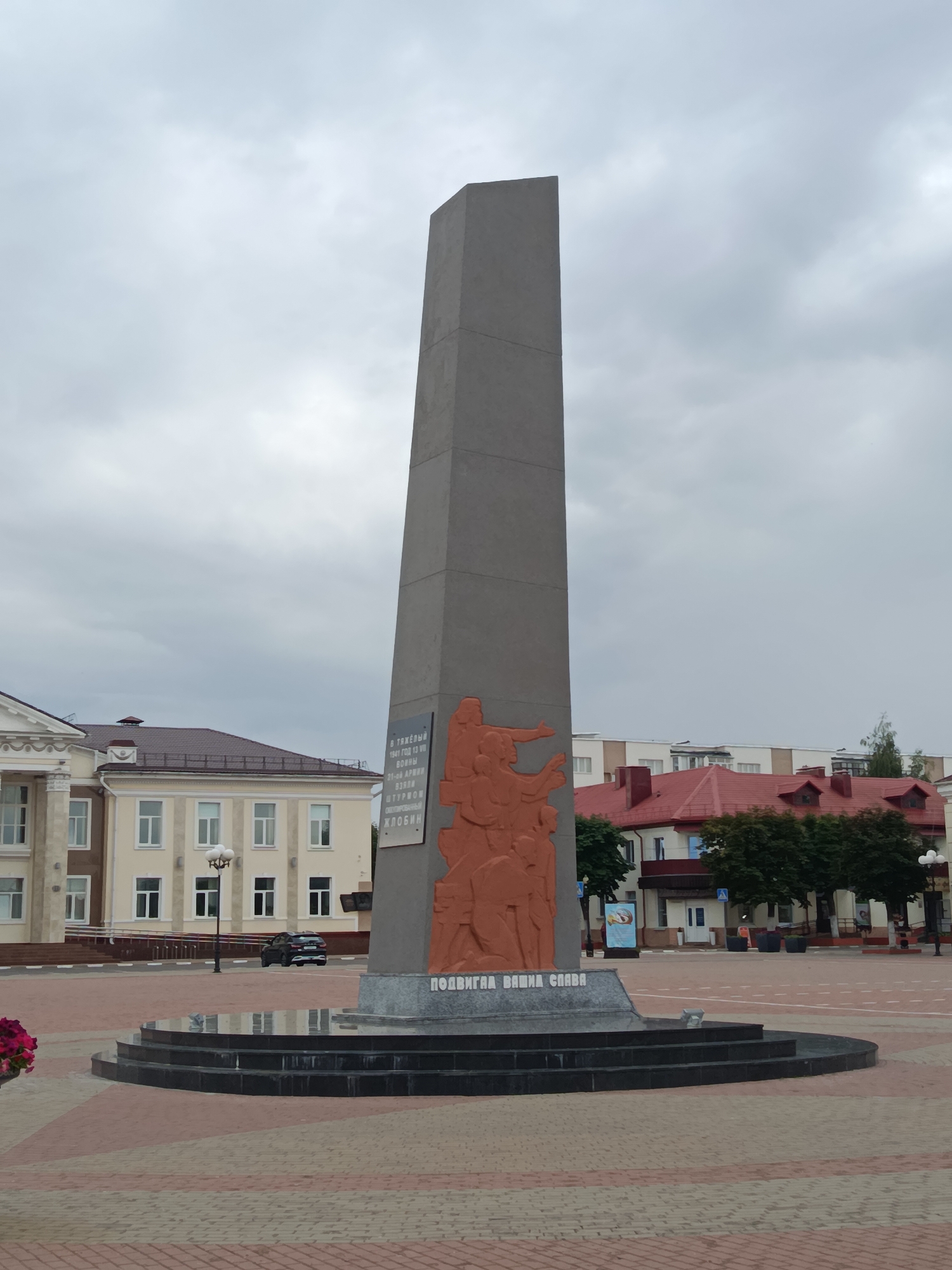 A gray obelisk stands tall in a public square paved with red and beige tiles. The obelisk features an orange bas-relief depicting a group of figures in motion. Below the bas-relief, text in Cyrillic reads "ПОДВИГ ВАША СЛАВА" (Your feat is glory). To the left of the obelisk is a light-colored, multi-story building with white-framed windows and columns. To the right and behind, red-roofed buildings and trees are visible. A black SUV is parked on the paved area in front of the building to the left. The sky is overcast.