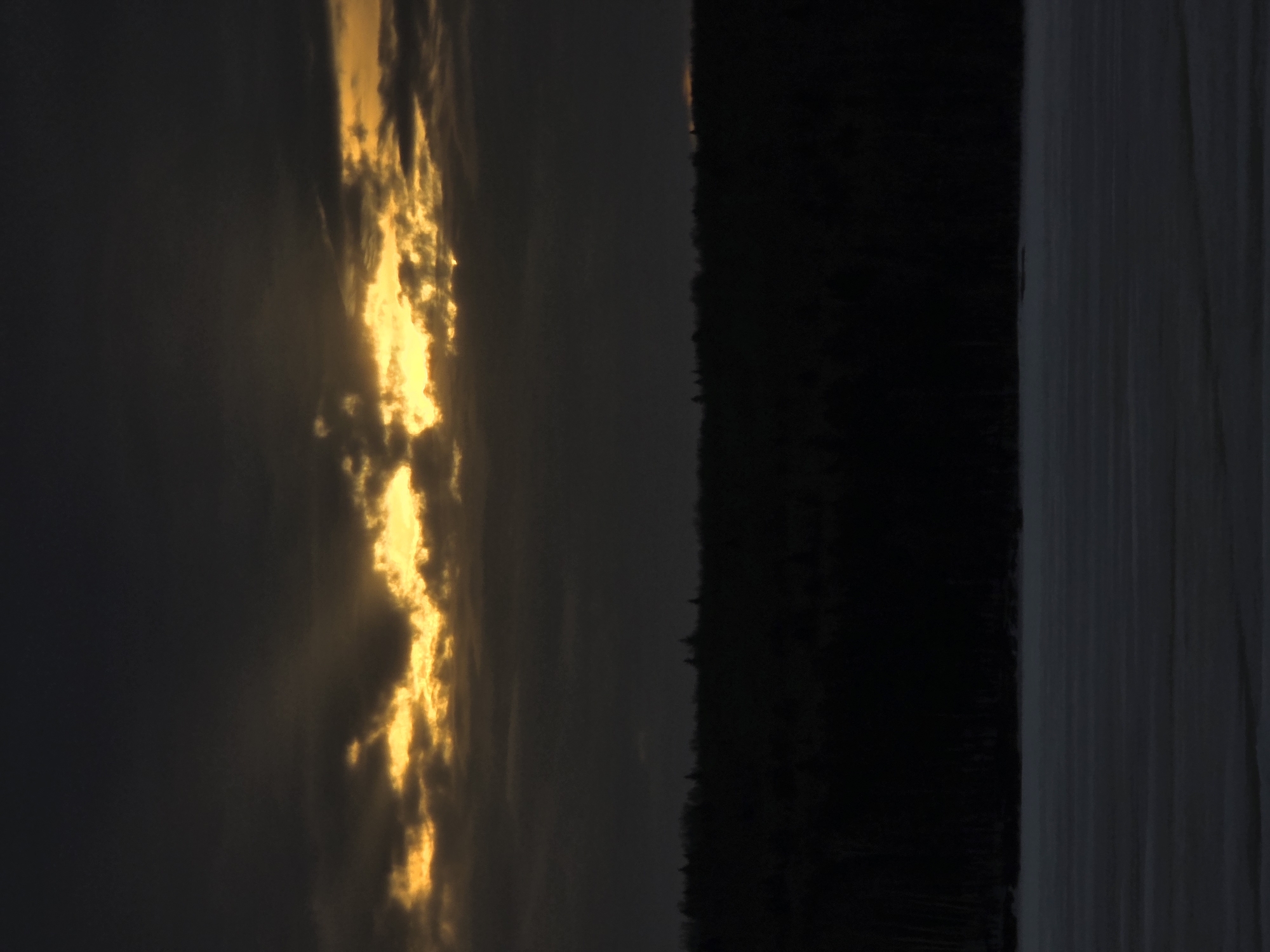 This photograph depicts a landscape in Chaikovsky, Russia. The sky, on the left, features dark clouds with a vertical streak of golden light, likely from the setting or rising sun. Next to the sky is a dense, dark line of trees forming a forest. Adjacent to the forest is a body of water. The light on the water reflects the color of the sky, making it appear white. The overall image has a dark and somewhat gloomy mood.