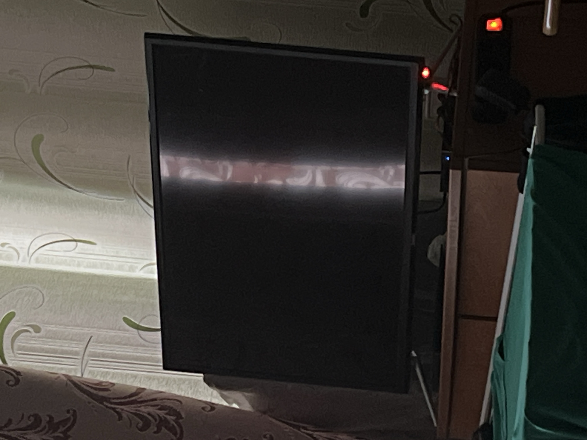 The image displays an indoor scene, likely a room such as a living room or bedroom. A dark, rectangular flat-screen monitor or television is centrally positioned, angled slightly. Its screen is black, but a bright, horizontal reflection spans across its middle, suggesting an external light source or window.
To the left of the monitor, the wall is covered with light-colored wallpaper featuring a repeating pattern of subtle green and beige swirling designs. In the foreground, below the monitor, a portion of a sofa or bed is visible, upholstered in a dark fabric with an intricate, lighter-colored floral or scrollwork pattern.
To the right of the monitor, the background is darker. A section of wooden furniture, possibly a shelf or cabinet, can be seen. Several electronic components are hinted at by a prominent glowing red indicator light in the upper right, another smaller red light below it, and a faint blue light, along with visible cables. A vertical strip of green fabric, possibly a curtain or part of a piece of clothing/storage, is present on the far right.
The overall lighting suggests a somewhat dim room, with the bright reflection on the screen being a notable contrast. No people are present, and there is no visible text. The image captures a static interior setting.