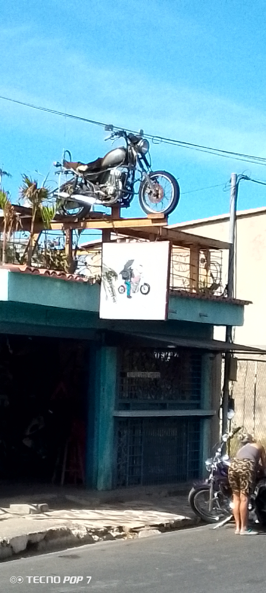 The image shows a street scene in Altagracia, Venezuela. A motorcycle is displayed on a platform built on top of a single-story building, with a sign hanging underneath. A second motorcycle is parked on the street in front of the building, where a person is interacting with it. The sky is clear and blue. The building appears to be a shop or garage. The sidewalk is broken and uneven.