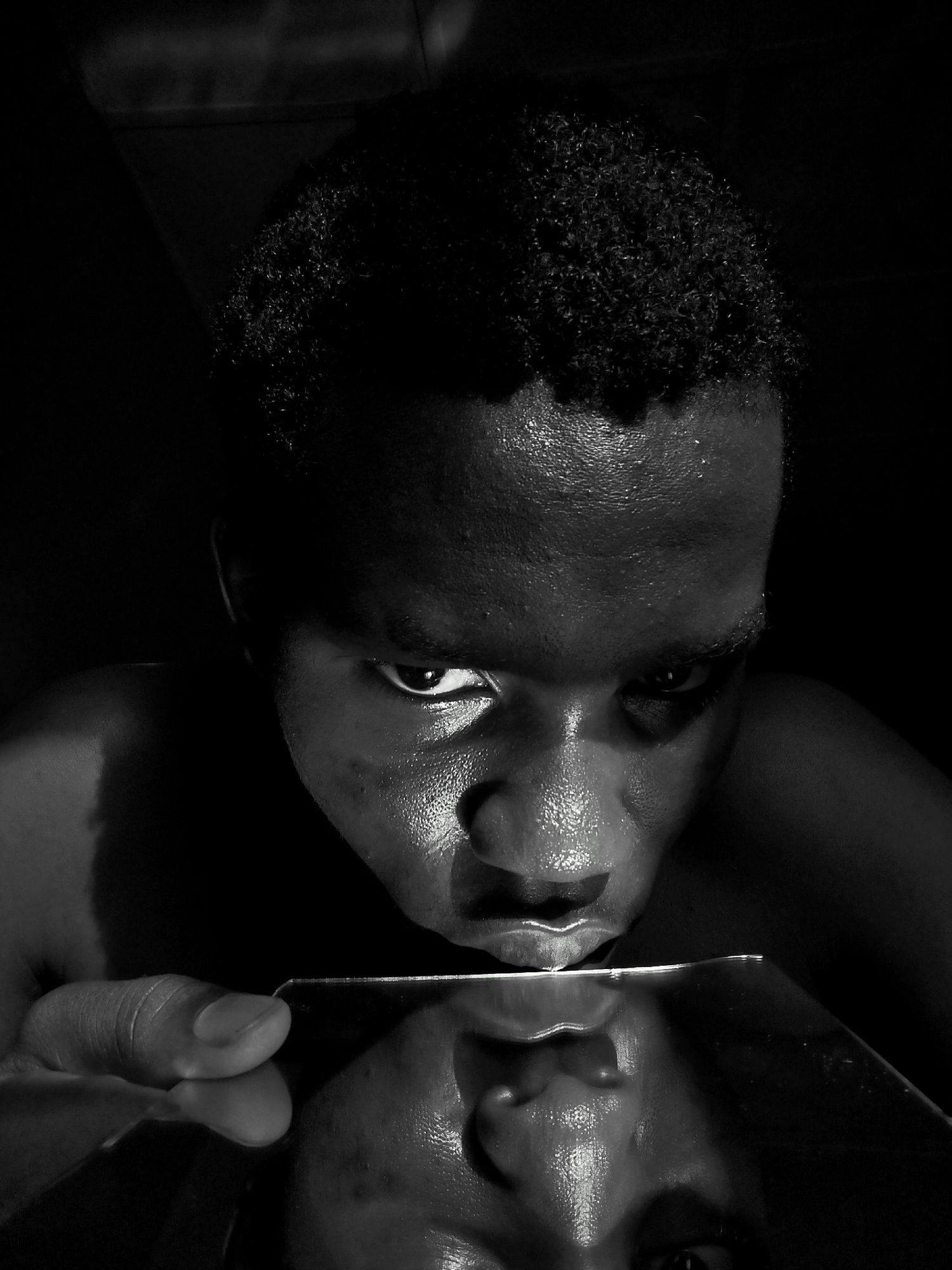 A black and white close-up shot of a person with short, curly hair gazing down at a reflective surface, possibly a mirror. The person's face is highly illuminated, emphasizing the texture of their skin and facial features. The reflective surface shows a blurred reflection of the lower part of the person's face, creating a double image effect. The hand holding the reflective surface is visible in the lower left corner of the image. The background is dark and largely undefined. The location is Unknown, Unknown.