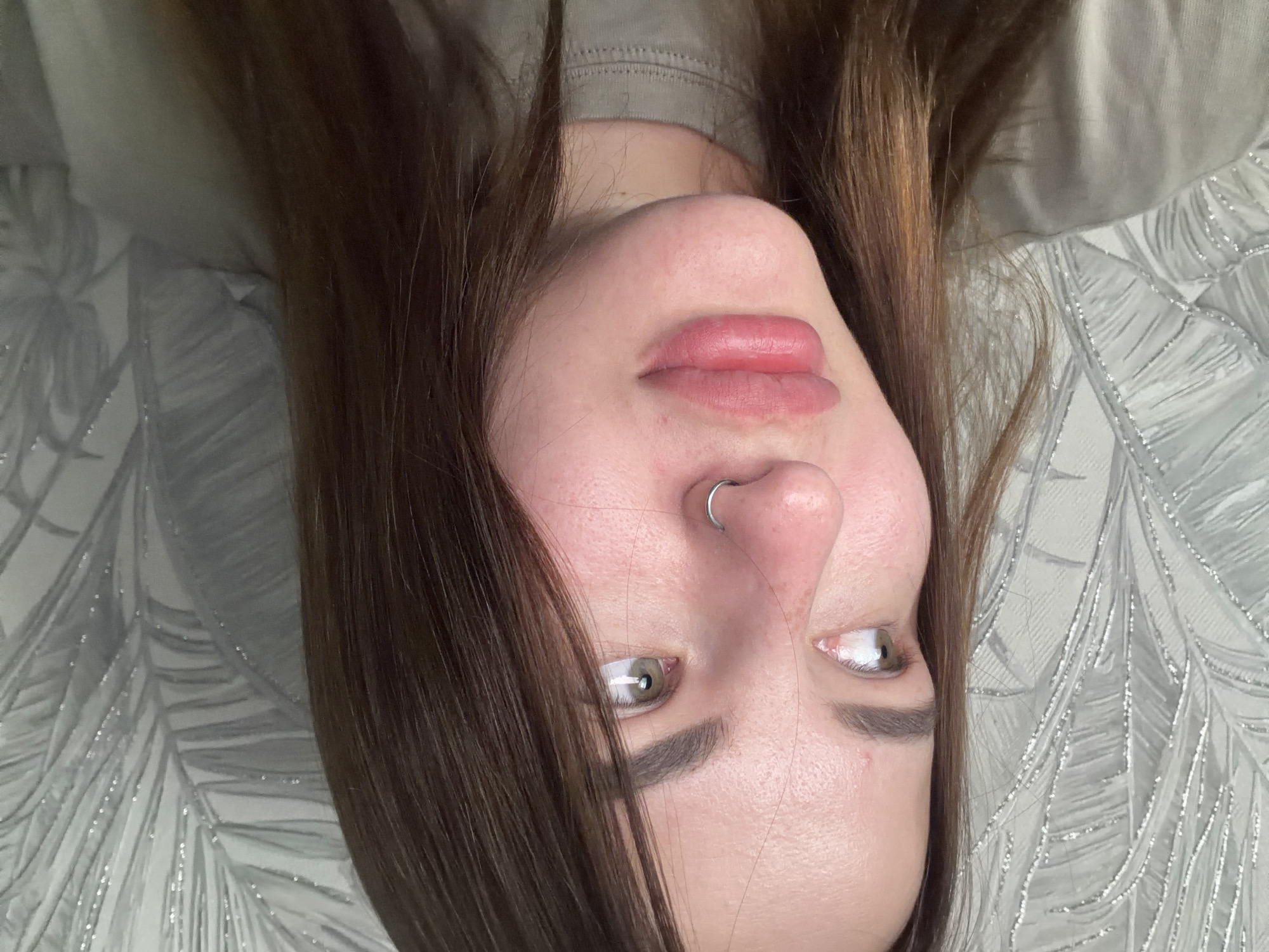 The image depicts a young woman with fair skin, brown hair, and green eyes, positioned upside down in the frame. She has a nose ring on her left nostril and defined eyebrows. Her lips are full and pink. She is lying on a gray and white patterned surface. A portion of what appears to be a light brown or gray shirt is visible at the top of the frame. The location or relevance to Novopokrovskaya, Russia, is not evident from the image.