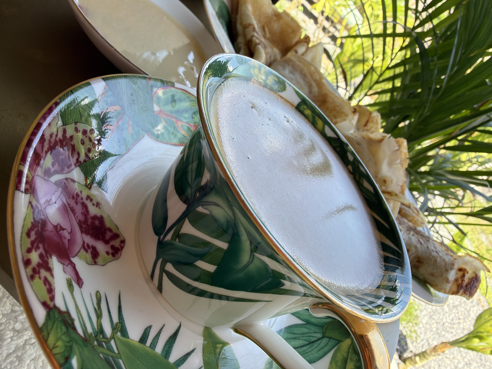 A close-up, eye-level shot focuses on a cup of coffee with a foamy top, placed on a saucer with a tropical leaf and flower pattern. The cup and saucer are white with green leaves and pink and purple flowers. A gold trim accents the cup's rim and handle. Behind the cup, a plate holds several rolled crepes. To the left of the cup, a small dish contains a light-colored liquid, possibly a sauce or cream. The background is blurred, showing lush green foliage, indicative of an outdoor setting, possibly a garden or patio in Ko Samui, Thailand. Sunlight filters through the leaves, creating bright spots. The overall scene suggests a leisurely breakfast or snack in a tropical environment.