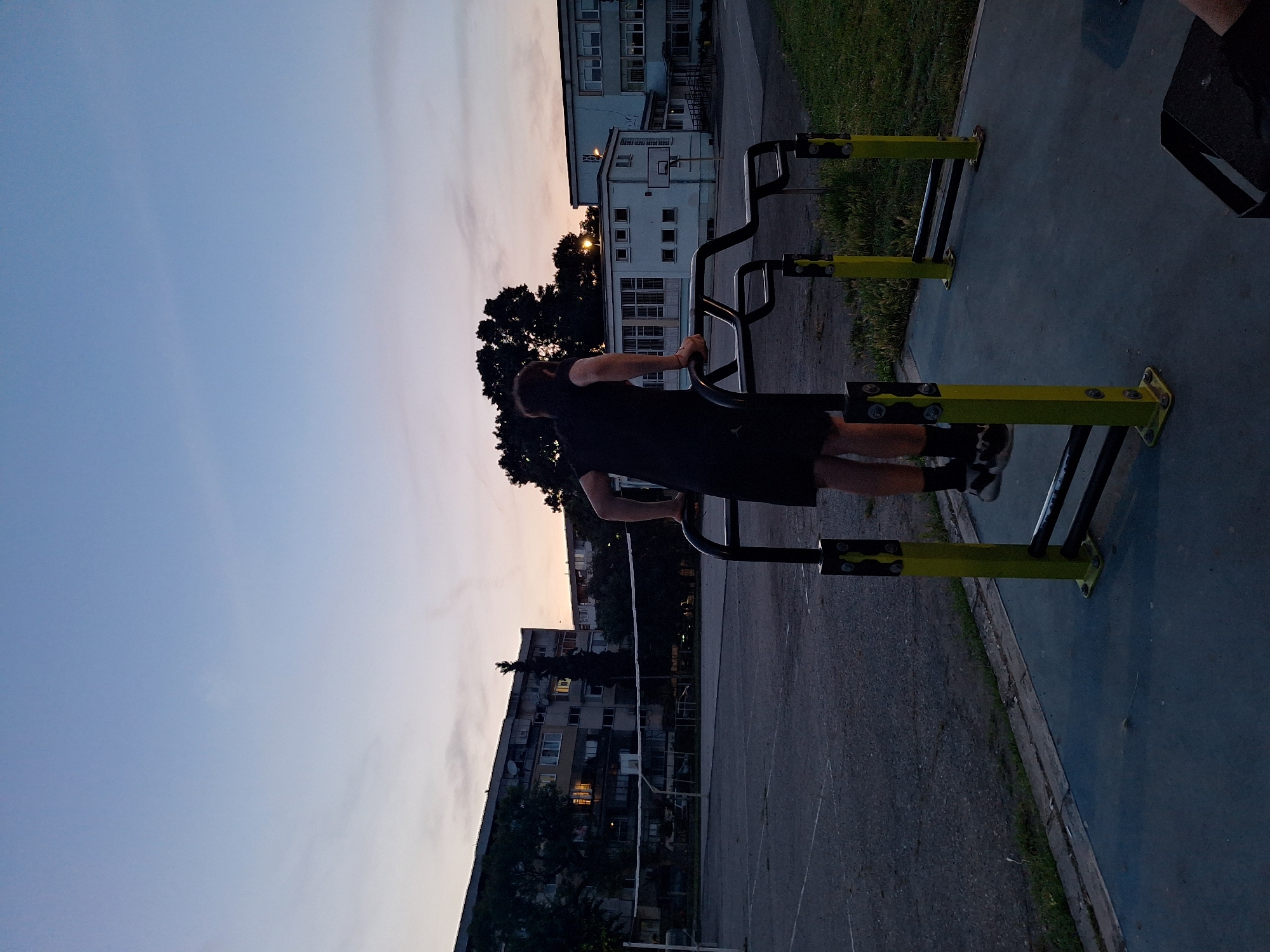 A person is doing pull-ups on a street workout gym in the city of Varna, Bulgaria, at dusk. The sky is a soft blue with hints of orange near the horizon, suggesting the sun has recently set. The person, wearing a black t-shirt and shorts, is positioned in the center of the frame, facing away from the camera, their hands gripping the parallel bars of the gym equipment. The gym has yellow and black supports. In the background, there are apartment buildings and trees, characteristic of a residential area. A volleyball net is visible in the middle ground, indicating a sports court. The ground is asphalt and concrete, with some patches of grass. A black bag is placed on the ground in the lower right corner of the image. The overall impression is one of a peaceful evening in an urban setting.