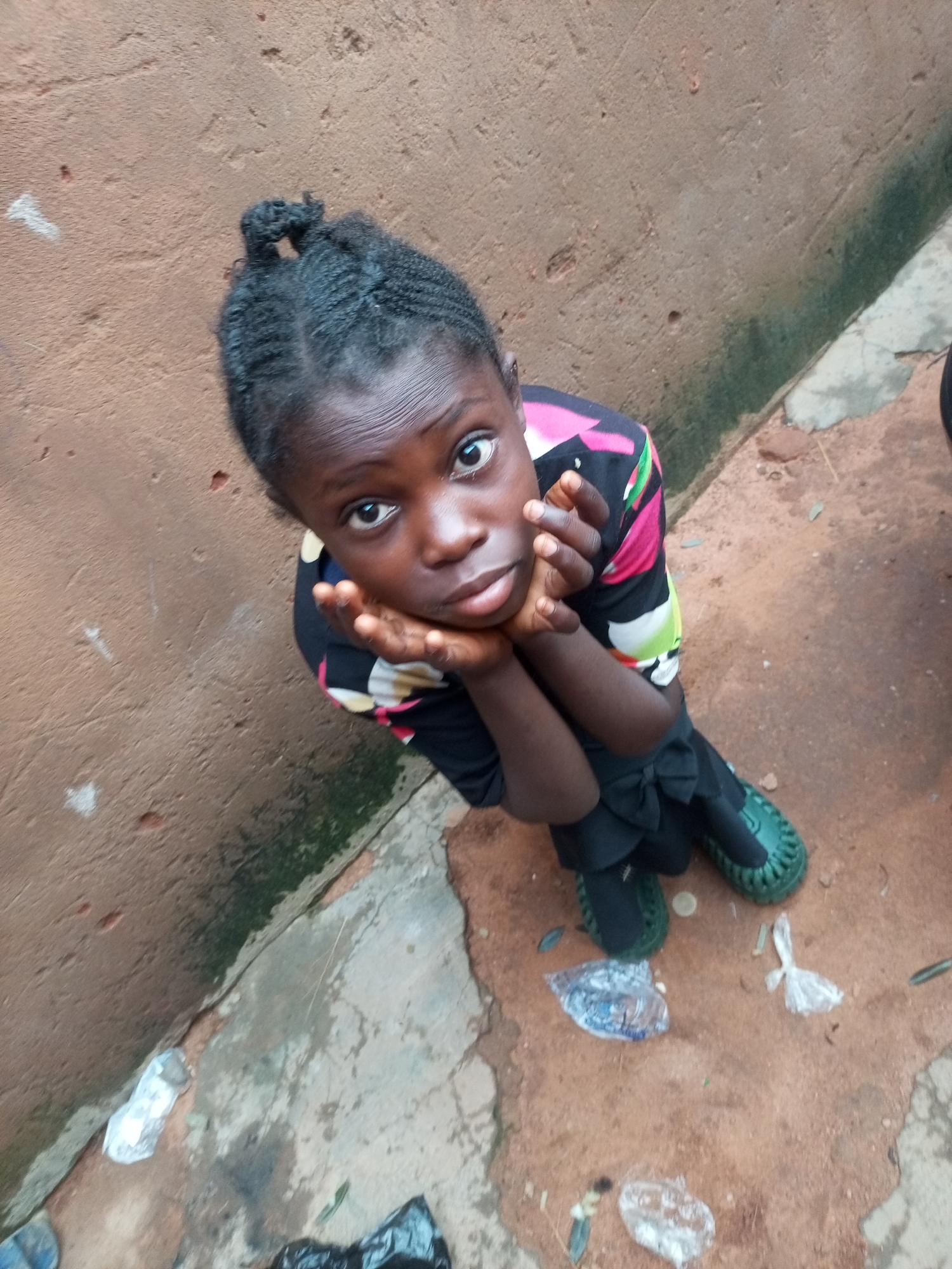 A young Black girl with her hair braided into a top knot is sitting on a dusty, cracked concrete ground next to a brown, rough-textured wall. She is looking up at the camera with her hands clasped under her chin, her expression curious. She is wearing a black shirt with a colorful floral print of pink, yellow, and green flowers. Her legs are crossed, and she is wearing dark green shoes. The ground around her is littered with small pieces of white and clear plastic, as well as some dry leaves. The scene suggests an outdoor environment, possibly in an alley or a less-maintained area. The lighting suggests it is daytime.