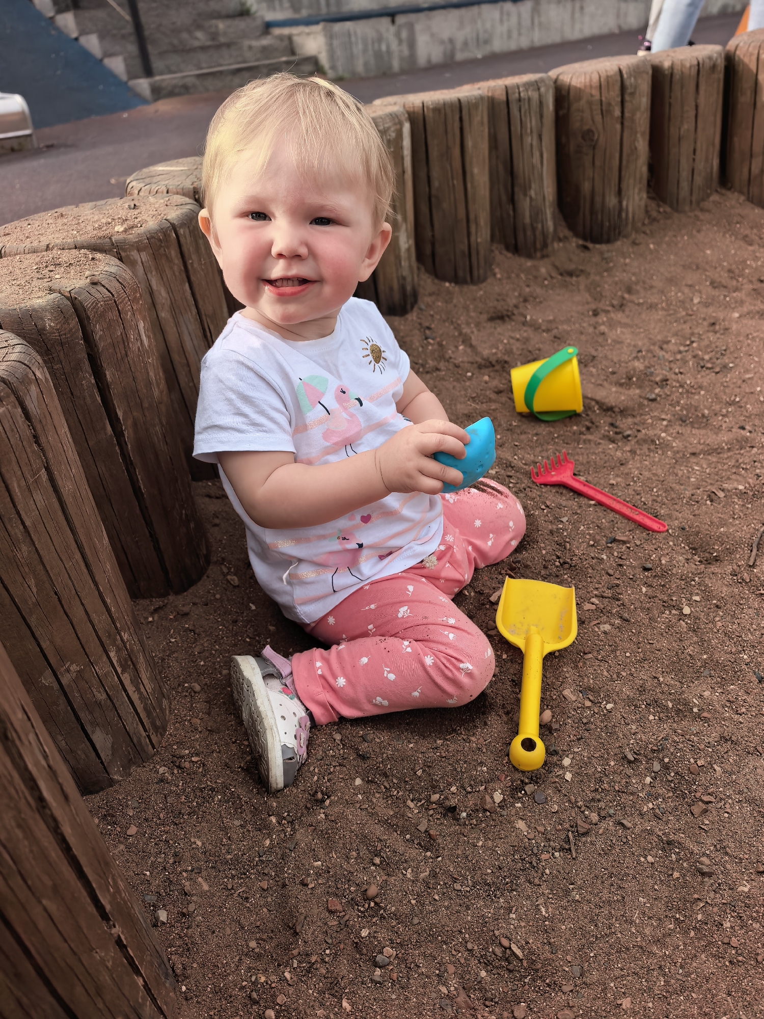 A young Caucasian toddler with blonde hair and blue eyes is sitting in a sandbox in Sterlitamak, Russia. The toddler is wearing a white t-shirt with a flamingo design and pink pants with white flower patterns. The child is holding a blue toy cup in their right hand and looking towards the camera with a smile.
In the sandbox, there are several toys, including a yellow shovel, a red rake, and a yellow bucket with a green handle. The sandbox is surrounded by a wooden border made of logs. The ground is covered in dirt and small stones. In the background, there are concrete stairs and a glimpse of what appears to be a playground or park area. The lighting suggests it is daytime, possibly late afternoon due to the warm tones. The overall scene depicts a child playing in a sandbox with toys. There is no visible text in the image.