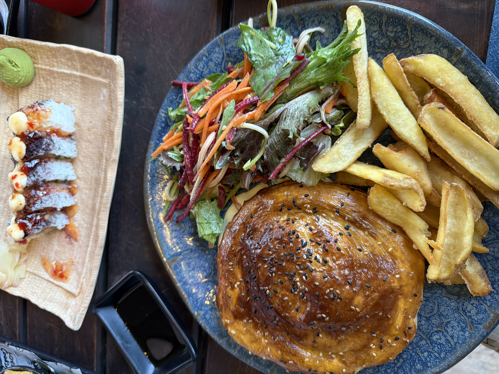 On a dark wooden table, a rectangular beige plate holds four pieces of sushi topped with what appears to be eel and a dollop of sauce, along with a scoop of wasabi and some pickled ginger. Next to it, a circular blue plate is laden with a meal consisting of a golden-brown baked pastry sprinkled with sesame seeds, a side of thick-cut fries, and a salad of mixed greens, shredded carrots, and beets. A small black dish with dark sauce sits between the two plates. The scene is a close-up shot of food served in a restaurant setting, likely in Varna, Bulgaria. The presence of sushi suggests a fusion cuisine or a diverse menu offering.