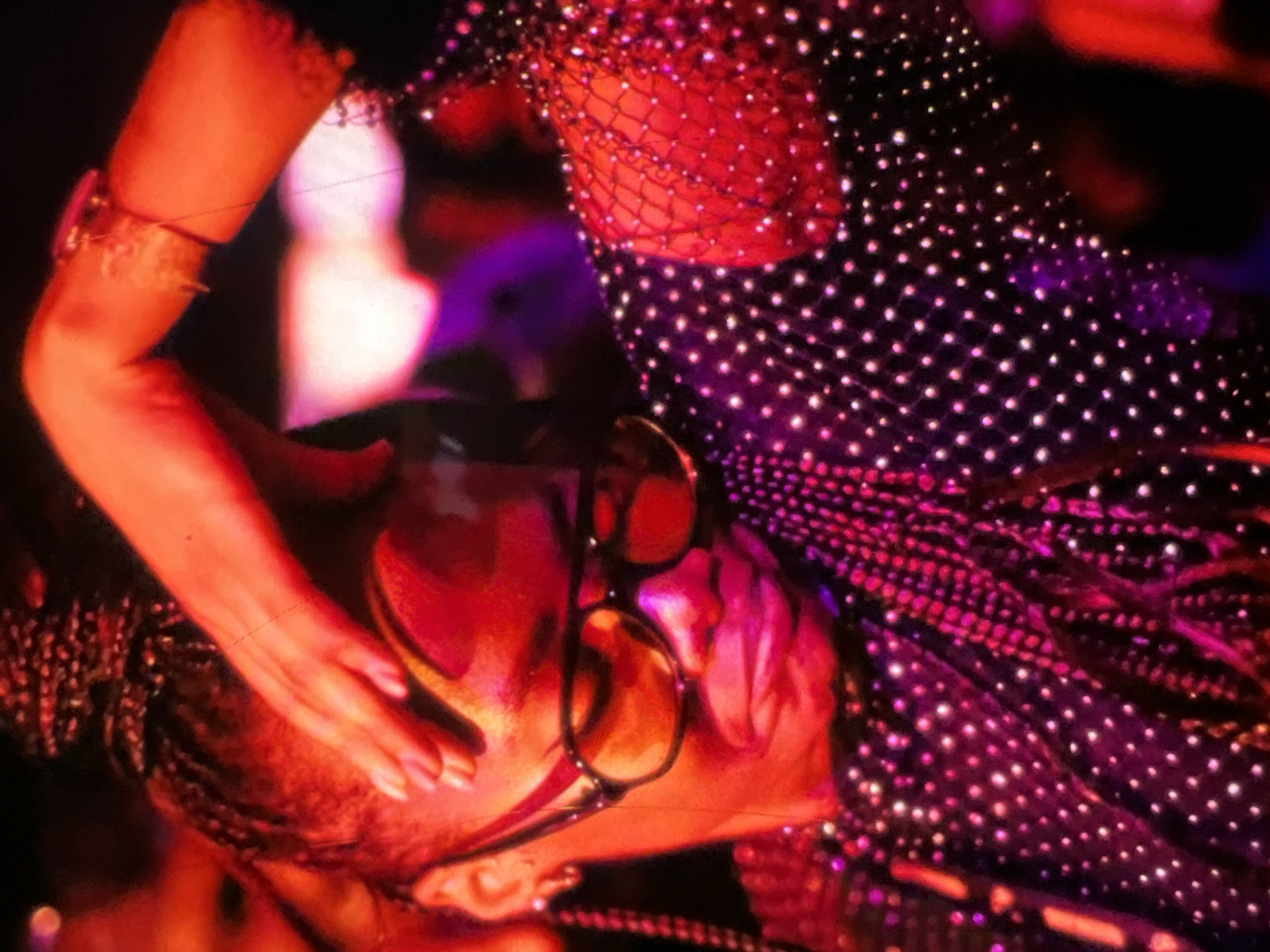The image shows a close-up, dimly lit, and heavily saturated photograph, dominated by red and purple hues, of a person. The subject is wearing dark-rimmed glasses and a garment adorned with numerous small, reflective beads or sequins, creating a sparkling effect. The person's right hand is raised to their forehead, obscuring part of their face. Their hair appears to be styled in braids, pulled back from their face. The background is indistinct, with areas of blurred light and dark, suggesting an indoor setting possibly a concert or club. There is no identifiable indication of the location being Lagos, Nigeria.