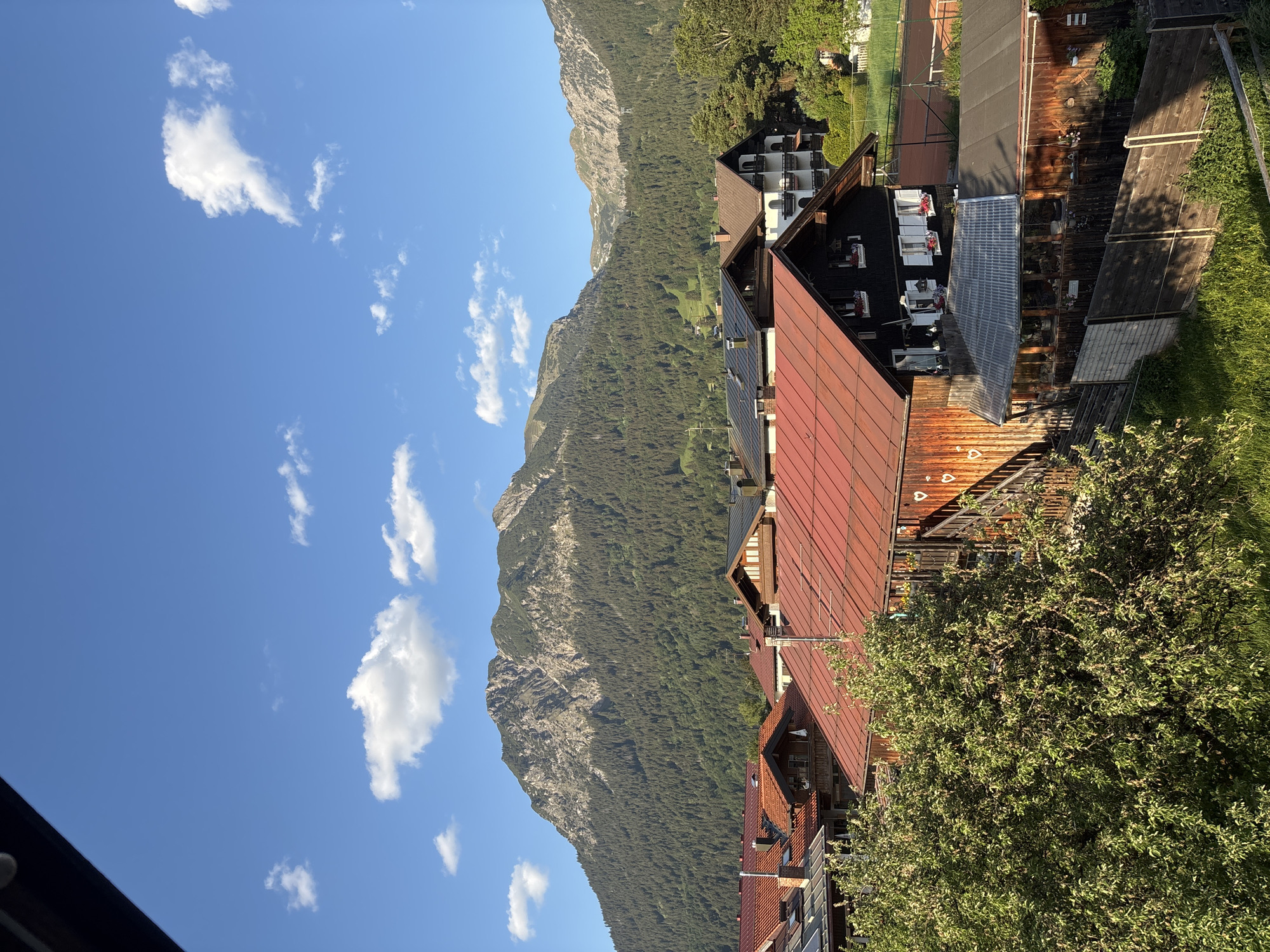 The image depicts a scenic view in Oberstdorf, Germany, looking towards a mountain range under a blue sky with scattered clouds. In the foreground are the rooftops of several buildings, some with red tiles and others with solar panels. There's also a tennis court visible, with lush greenery surrounding the structures, suggesting a blend of residential and recreational areas amidst the natural landscape. The mountainous backdrop is characterized by dense forest cover, enhancing the picturesque setting of the Bavarian Alps.