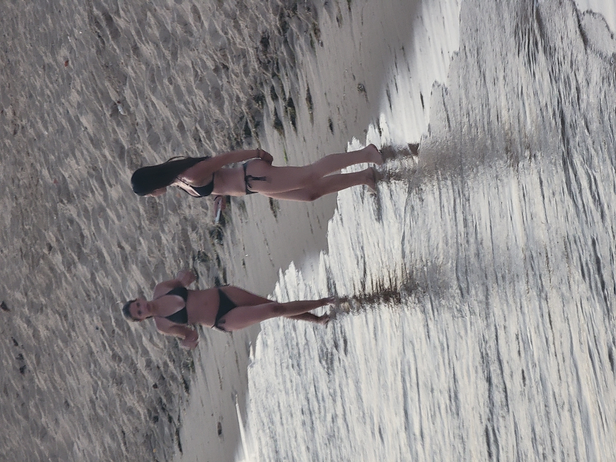 Two women stand at the edge of the ocean on a sandy beach. The woman on the left, facing the viewer, is wearing a dark bikini and has her arms slightly bent at the elbows with her hands open. The woman on the right, facing left, is also wearing a dark bikini and appears to be looking out towards the water. The sand behind them is textured with small ridges and appears dry. The water in the foreground is shallow and reflects the light, with small waves lapping at the shore. This scene is located on the Quarteira beach in Quarteira, Portugal.