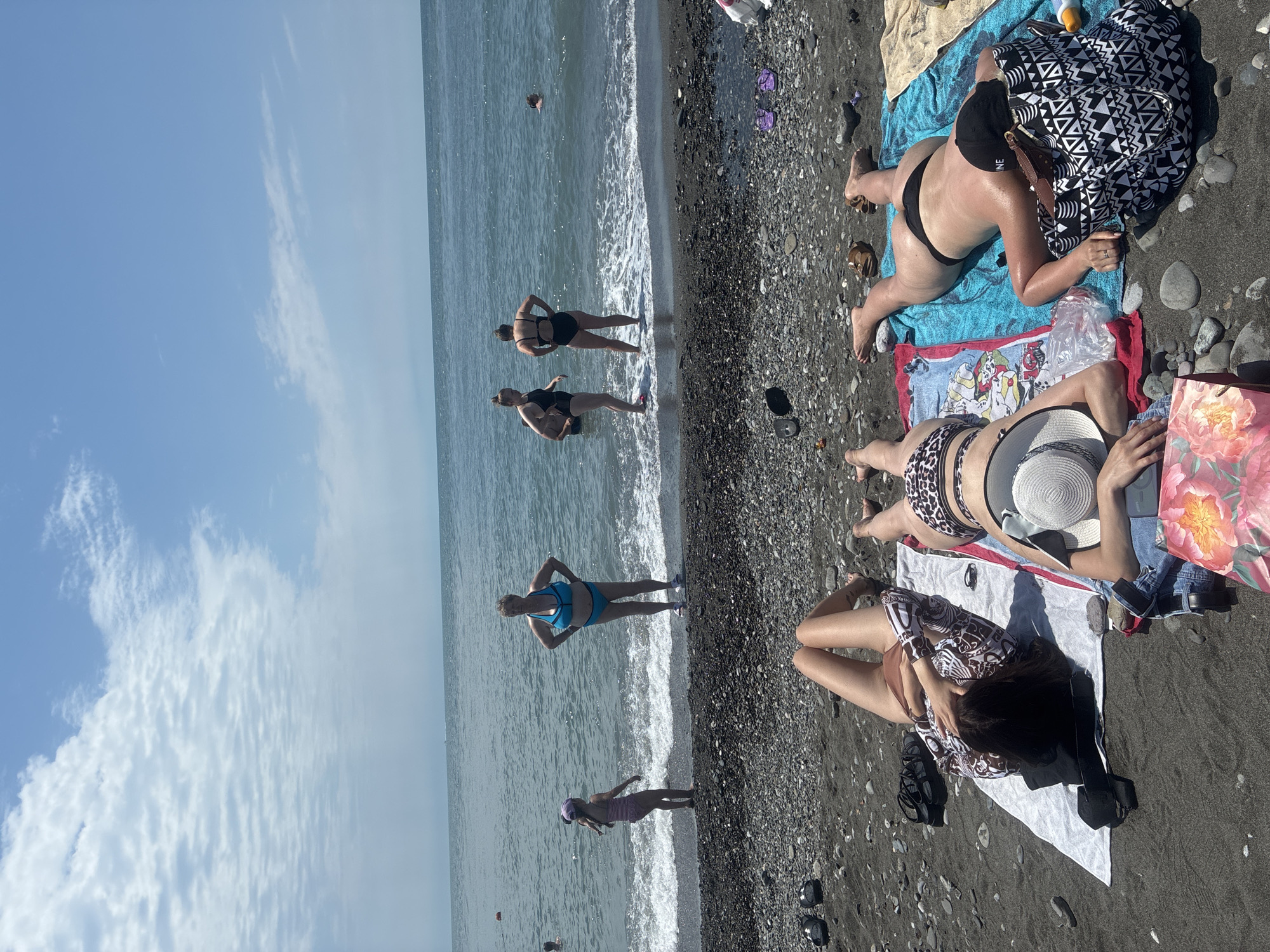 The image shows a group of people on a pebble beach in Sirius, Russia. The sky is partly cloudy with a clear horizon over the sea. In the foreground, three women are lying on towels on the beach, sunbathing. The woman on the far left is wearing a patterned top and brown bikini bottoms, lying on her back with her arm over her face. The woman next to her is wearing a leopard print bikini and a white sun hat, lying on her stomach. The woman on the far right is wearing a black bikini and a black cap, lying on her stomach with her head turned away. In the background, several other people are in the water, with two women standing at the edge of the water, and a child in a striped swimsuit standing near them. The beach is covered in dark pebbles, with some scattered stones and a few beach accessories.