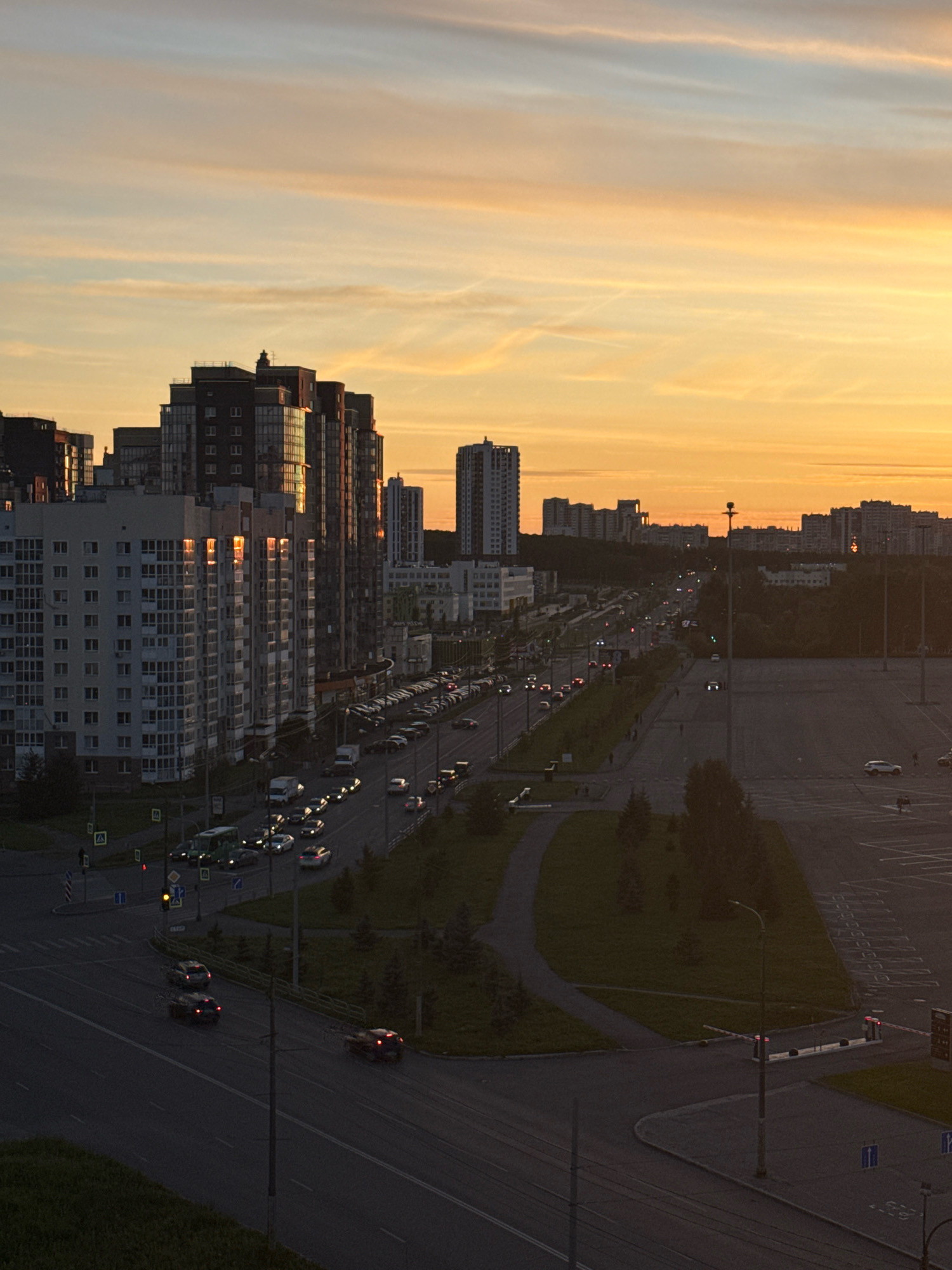 This is an aerial view of a cityscape at sunset, featuring a network of roads, traffic, and several multi-story buildings. The sky is a vibrant orange and yellow, indicating the sun is low on the horizon.
The foreground shows a dark asphalt road with several cars, some with their brake lights illuminated, suggesting they are in motion or stopped. A pedestrian walkway with grass and trees lines the side of the road.
Further back, a multi-lane road is visible, with a significant amount of traffic including cars and a bus. The buildings in the midground are modern, residential or office towers, with glass facades that reflect the warm colors of the sunset. Some of these buildings are quite tall.
In the background, more buildings form a skyline, some appearing to be apartment blocks or commercial structures. A large, empty parking lot is present to the right of the image, with painted parking spaces visible. There are no discernible people in the image, other than a few distant figures walking on a path in the midground.
This scene is likely located in the residential area of the city of Chelyabinsk, Russia, possibly near one of the newer developed districts with high-rise apartment complexes and surrounding infrastructure. The time of day is either sunrise or sunset.