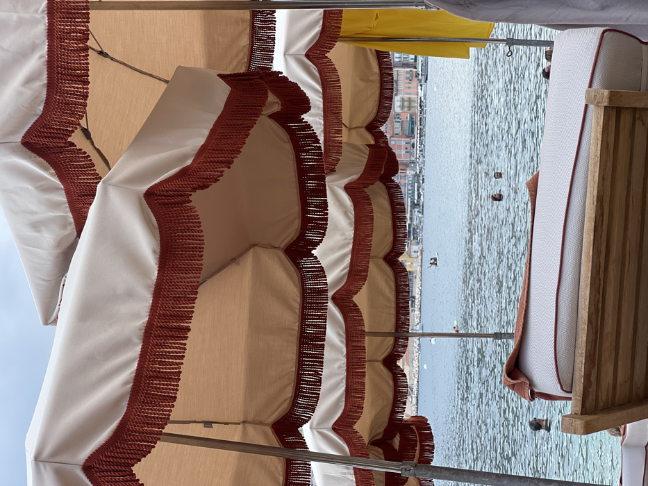 The image showcases a row of beach umbrellas with a white and tan color scheme, adorned with brown fringe along the edges. The umbrellas offer shade over a body of water, in which a few boats and buoys are floating. The water stretches to the horizon, where buildings are visible. A wooden beach chair with a white cushion is partially in view, adding a sense of relaxation.