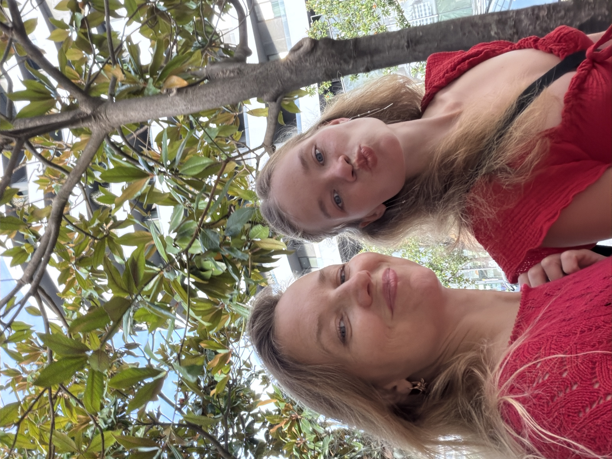 The image shows a close-up, selfie-style shot of two women outdoors in Budva, Montenegro, with a tree and foliage in the foreground, and a modern building in the background. The woman on the left, in the foreground, has blonde hair and is smiling directly at the camera. She is wearing a red, patterned top. The woman on the right is slightly behind her, also in the foreground, with blonde hair and is puckering her lips towards the camera. She is also wearing a red top. The tree branches and leaves are prominent in the upper portion of the image, obscuring parts of the sky and building. The building in the background appears to be a multi-story structure with white and glass elements. The overall lighting suggests it is daytime.