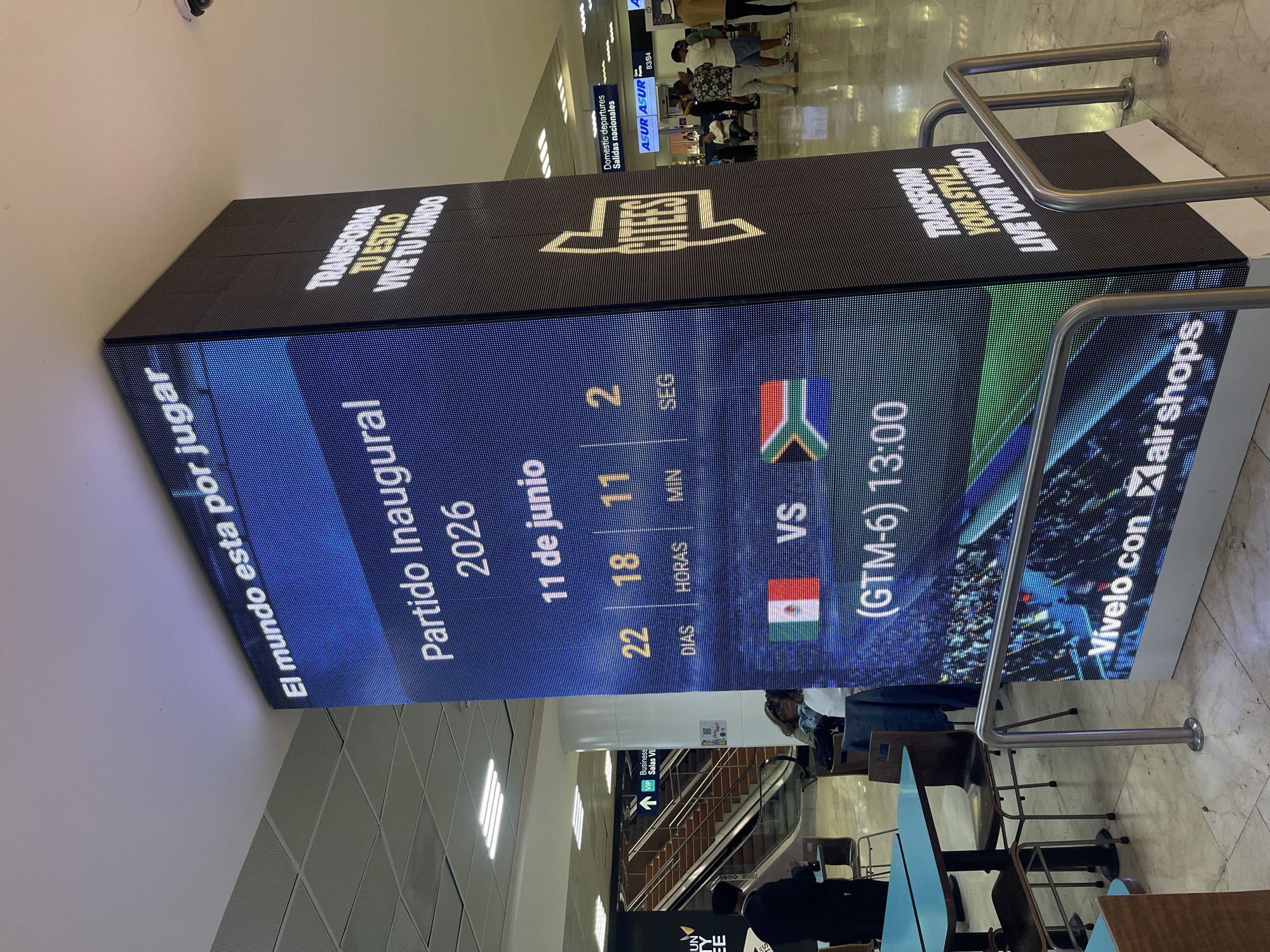 The image shows a large LED display screen in what appears to be an airport. The screen displays information about the inaugural game of the 2026 soccer (football) tournament, scheduled for June 11, 2026. The display indicates a match between Mexico and South Africa, and the countdown timer is showing 22 days, 18 hours, 11 minutes, and 2 seconds. The time of the match is listed as 13:00 in the (GTM-6) time zone. The logo for ASUR, the airport group, is visible, as well as "Vívelo con airshops" (Live it with airshops). Based on the signs above that show "Salidas Nacionales (Domestic Departures)" in the photograph, this appears to be inside an airport terminal. Additionally, a sign on the display states, "Transforma tu estilo, Vive tu mundo (Transform your style, Live your world)". The presence of an escalator and seating area further suggests an airport environment. The city of Cancún, Mexico is mentioned in the context, although not overtly visible in the image.