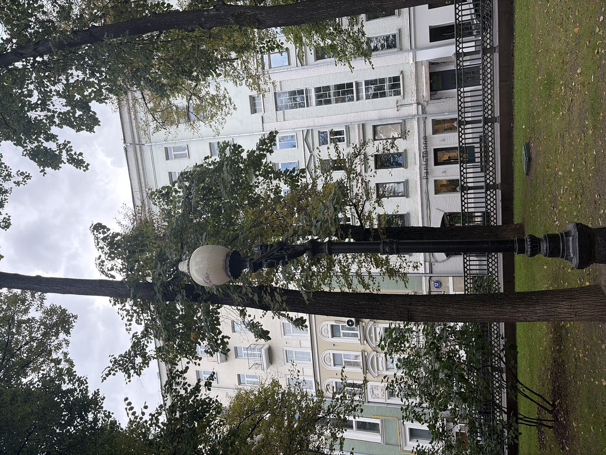 The image captures an overcast day in Moscow, Russia, with a focus on a tall, light-colored residential building partially obscured by trees. In the foreground, a vintage black lamppost stands prominently. The building exhibits a blend of architectural styles, with several floors featuring numerous windows of varying designs, some with balconies. The lower facade of the building appears to house shops, indicated by signage such as "Parisienne." A black metal fence lines the area in front of the building, separating it from a grassy area with scattered fallen leaves. The trees, with their green foliage, are a dominant feature, framing the scene and suggesting a park or courtyard setting within the city. The sky is cloudy, contributing to the subdued lighting of the photograph. A blue sign with the number "12" is visible on the building near the fence.