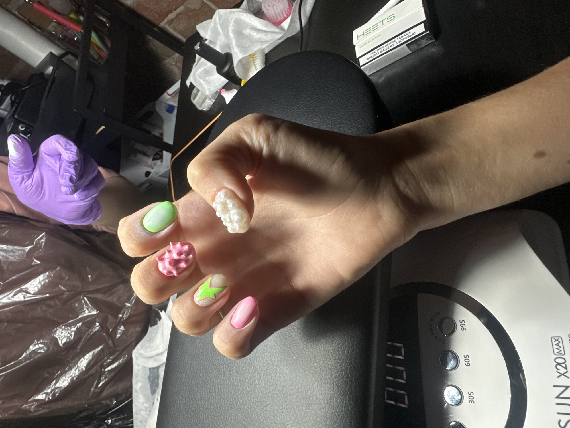 A close-up shot shows a person's hand with colorful, textured, and decorated fingernails. The hand is resting on a black surface, likely a table, next to a white LED nail lamp with digital display showing "000" and several timer settings labeled "30S," "60S," "99S," and "LOW." To the upper right, a pack of HEETS Green Selection cigarettes is visible, with Russian text on the pack that translates to "Smoking tobacco harms your health." A purple glove is on the person's left hand, which is partially visible in the background, suggesting a manicure or nail art session is in progress. The overall setting appears to be indoors, possibly a home or salon, with brickwork and shelves visible in the background. The lighting is artificial, typical of an indoor environment. The focus is on the hand and the detailed nail art, which includes various colors like pink, green, and white, with some nails featuring 3D embellishments.
