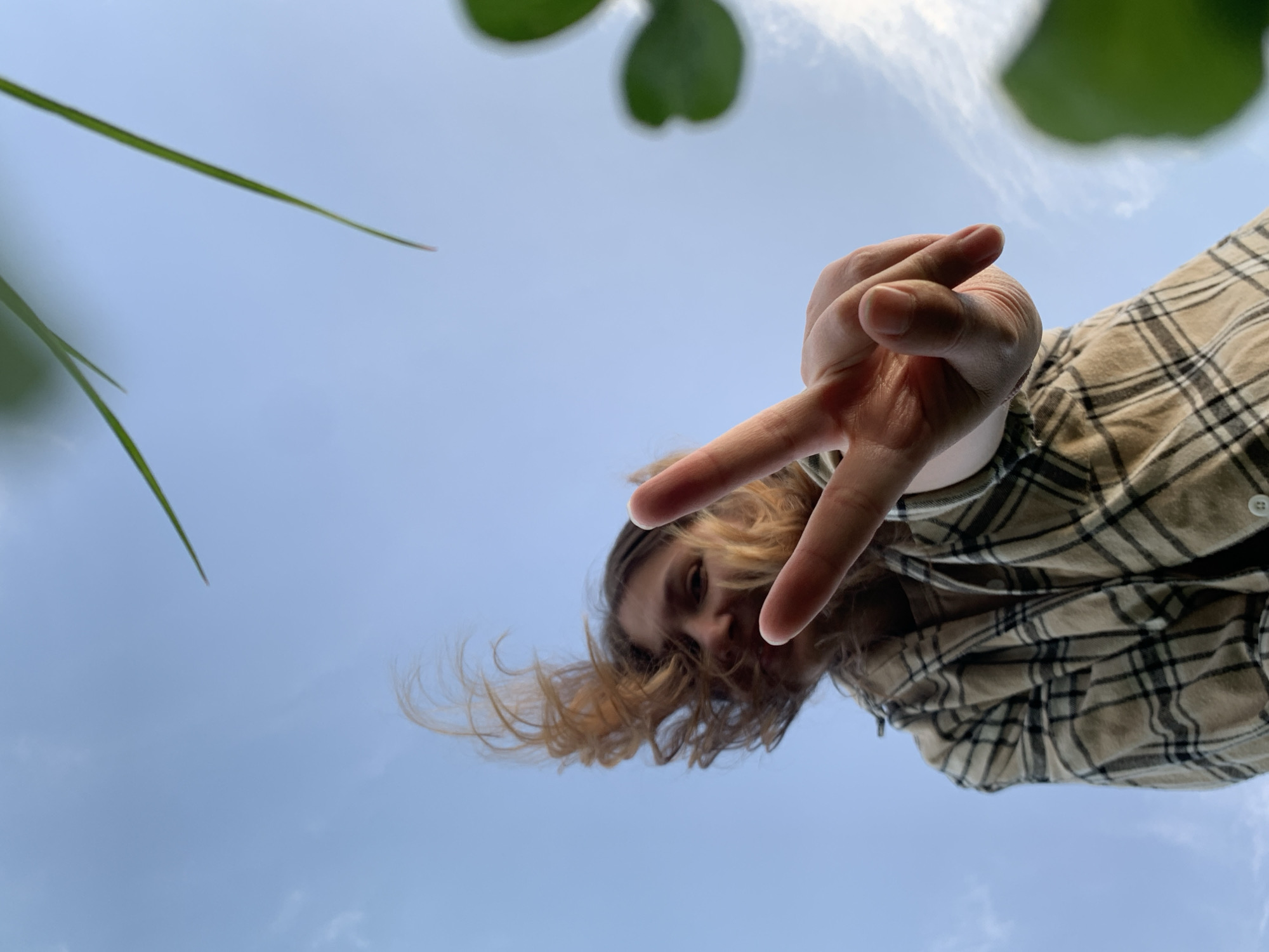 A low-angle shot of a person, possibly in Chaikovsky, Russia, against a sky background. The person is wearing a plaid shirt and making a peace sign with their hand. Hair is partially covering their face, which is only partly visible. Some greenery, possibly leaves or grass, is in the top left and right corners of the frame. The background is a light blue sky with some faint clouds. The specific location in Chaikovsky is not discernible from the image.