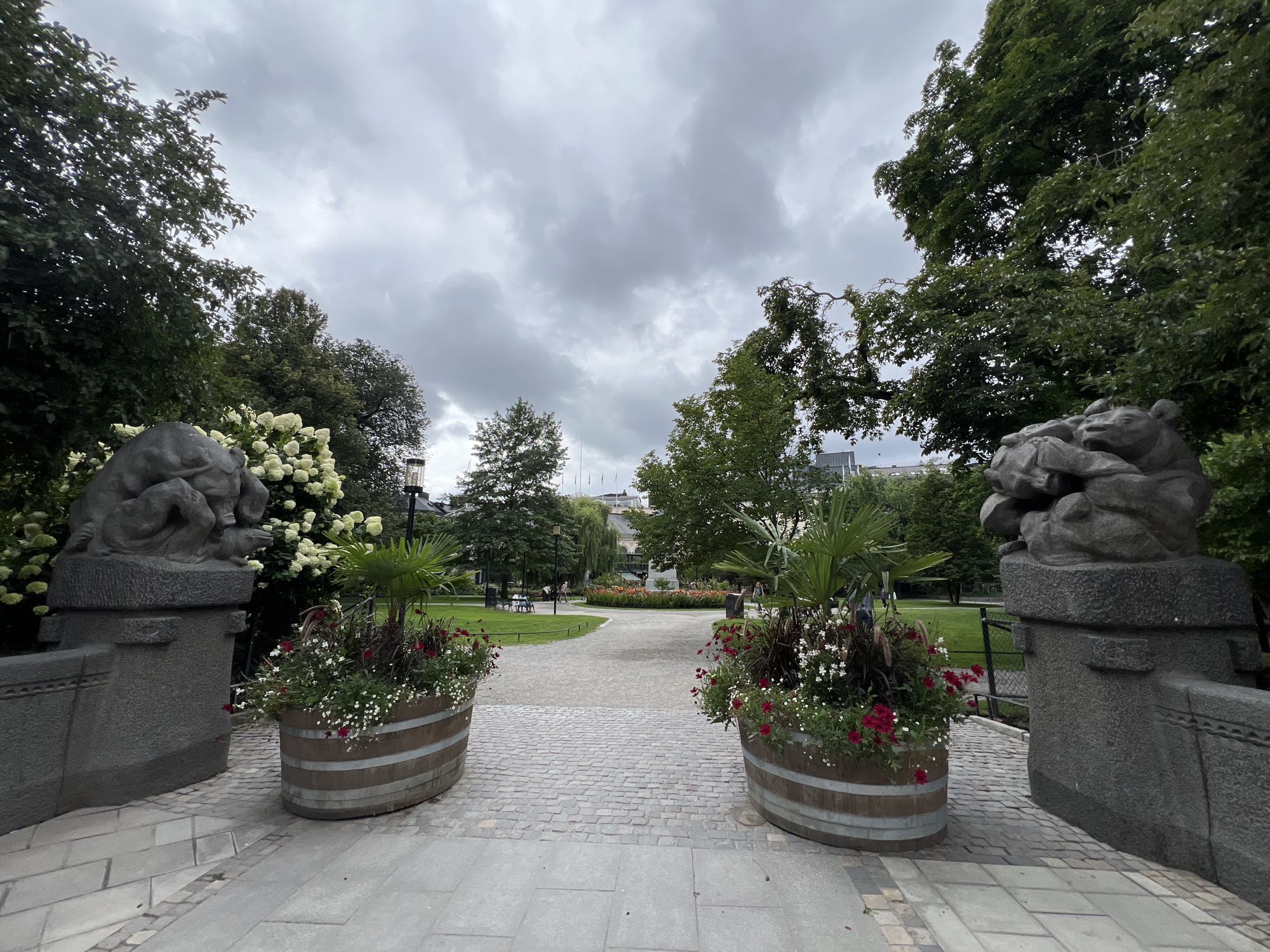 This image depicts the entrance to Kungsträdgården, a park in Stockholm, Sweden. The perspective is from a paved pathway leading into the park, with two large stone sculptures of lions flanking the entrance on either side. In front of each sculpture are large wooden planters filled with red and white flowers and greenery. Beyond the planters, a cobblestone path curves into the park, leading towards a grassy area with trees and a flower bed in the distance. Several people are visible in the park, some sitting on benches and others walking. The sky is overcast with dramatic clouds. The overall scene conveys a sense of a public park entrance on a cloudy day.