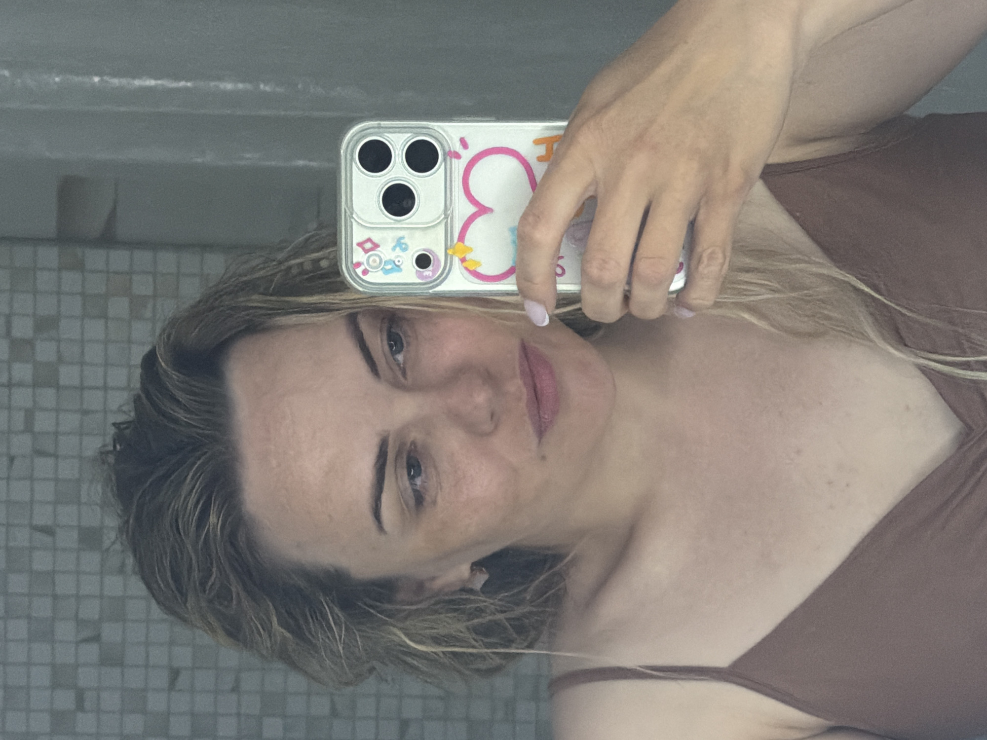 A woman is taking a selfie in a bathroom with tiled walls. She is holding an iPhone in a clear case with a cartoon design. She is wearing a brown tank top. Her hair is blonde and wavy. The photograph does not include any information about the location of the city of Votkinsk, Russia.
