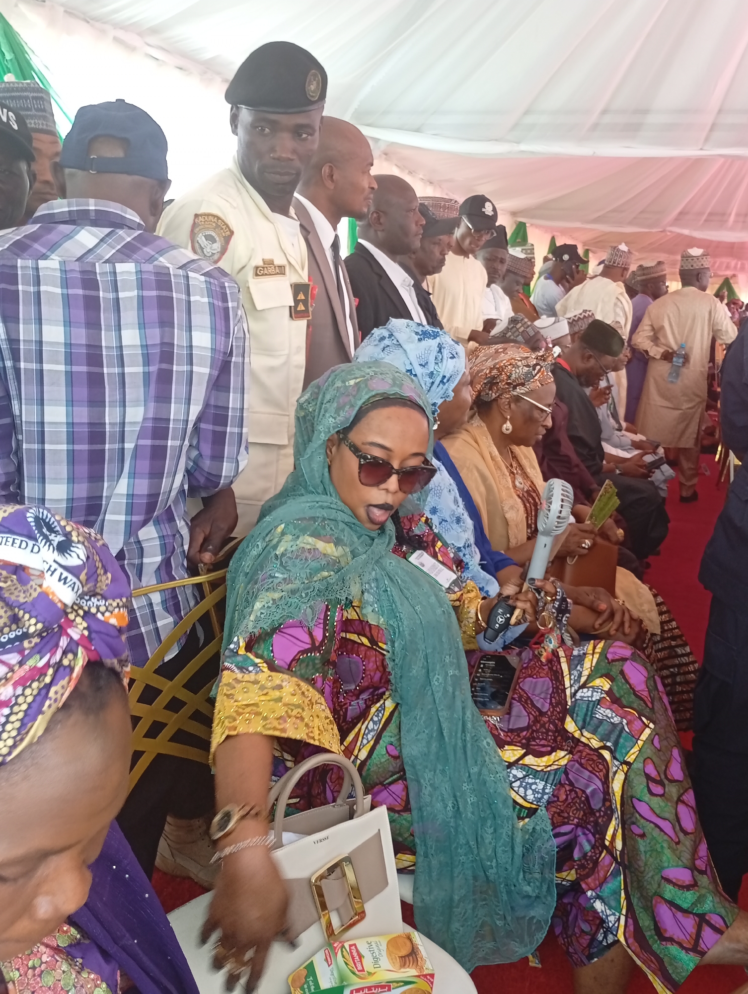 This media file captures an indoor gathering or event, likely a formal or community function, held under a large white tent with red carpeting in Unguwan Sanusi, Nigeria. The scene is populated by numerous attendees, predominantly adults, including both men and women.
In the foreground, several women are seated. A central figure wears a vibrantly patterned dress in shades of purple, green, yellow, and red, draped with a light green, lace-patterned hijab. She has dark sunglasses on and her mouth is slightly open, suggesting she might be speaking or reacting. On the table in front of her is a cream-colored handbag with "VERSSE" visible, and a box of "Britannia Digestive" biscuits, also labeled "بريتانيا" in Arabic. She holds a Mercedes-Benz key fob. To her right, another woman, also seated, wears a gold-colored outfit and head covering, and is holding a white portable fan.
Behind the seated women, a mix of men are standing and observing. A man in a light-colored uniform and a black beret is prominent; his uniform bears a patch reading "ADUNA STATE" and a name tag with "GARBA". He is looking towards the viewer. Other male attendees wear traditional Nigerian attire, including various caps and flowing garments, while some are in shirts, such as the man with his back to the camera wearing a purple and white checkered shirt. Partially visible text on clothing includes "TEED D" and "SH WA".
The overall impression is of an attentive audience at a public gathering, likely during daytime given the bright, indirect lighting within the tent.