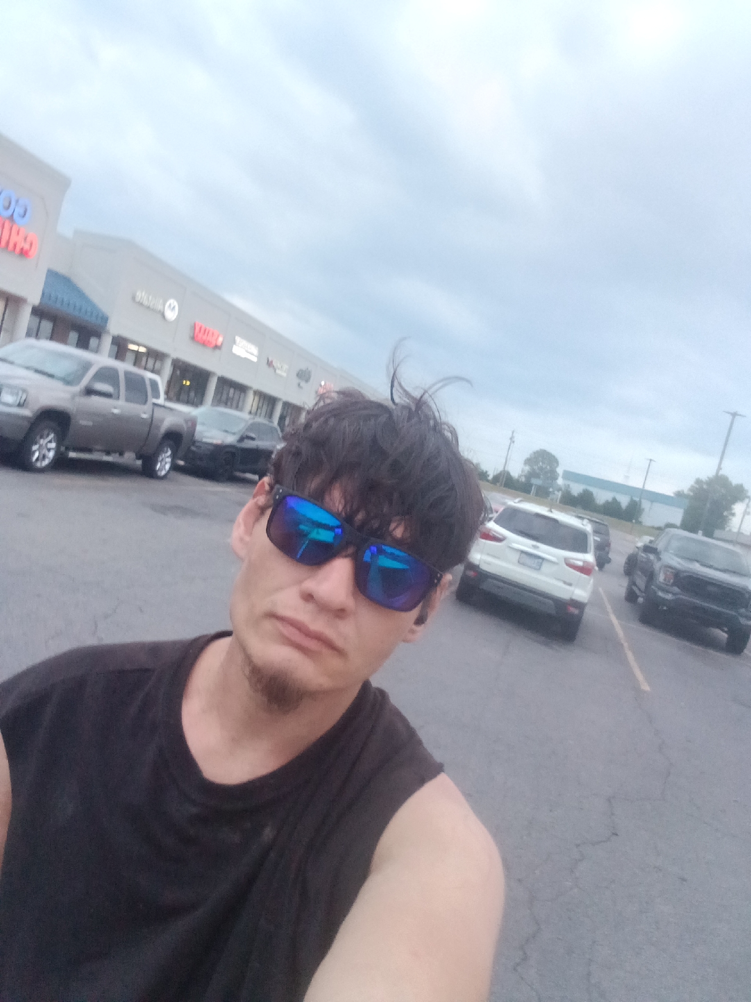 A selfie taken in a parking lot in Van Buren, Arkansas, USA. The individual is wearing blue-tinted sunglasses and a dark tank top. Behind the person, cars are parked in the lot, with a commercial building visible in the background. The sky is overcast.