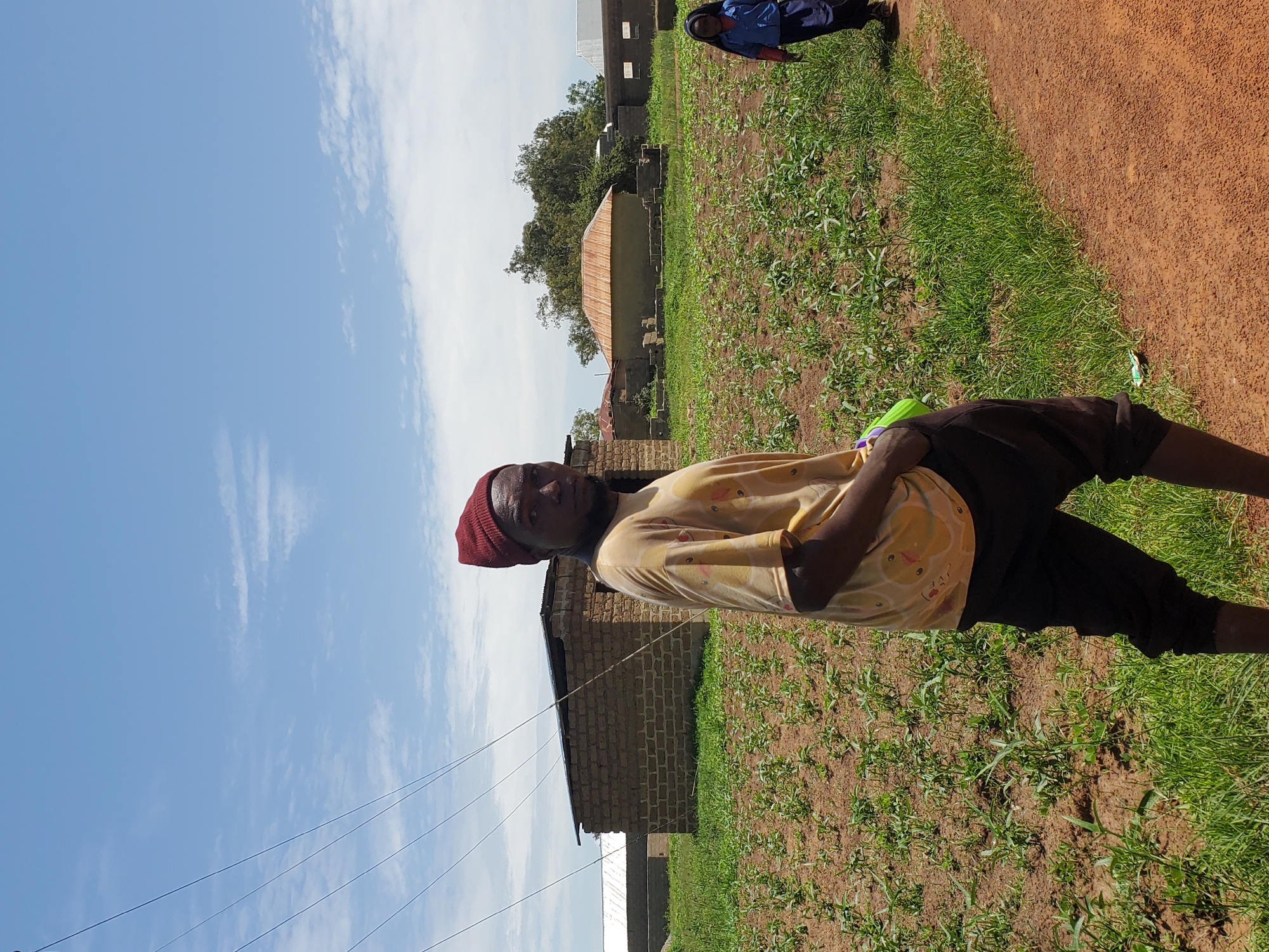 In Pankshin, Nigeria, a young man wearing a red beanie and a yellow t-shirt stands in the foreground, looking to the left of the camera. He is positioned on a dirt path next to a field of young crops. Behind him, a brick building with a corrugated metal roof stands under a blue sky with scattered clouds. Further in the background, another similar building and a round hut with a rusty metal roof are visible. A young girl in a blue shirt and dark skirt walks away from the camera on the dirt path to the right. The environment is rural, characterized by agricultural land and simple structures.