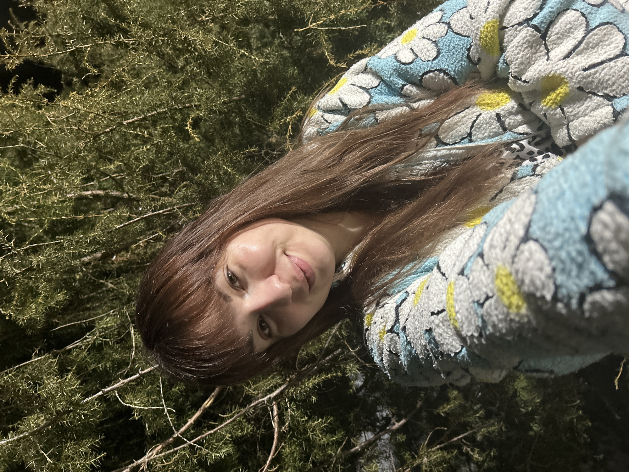 The image displays a selfie of a female in the city of Фоки, Russia. The subject, a woman with long, brown hair styled with bangs, is positioned in the foreground, facing the camera with a slight smile. She exhibits fair skin and clear facial features. She is wearing a textured, fuzzy garment, patterned with white daisies outlined in black, featuring light blue petals and yellow centers against a darker blue base. Her right arm is visible in the lower right portion of the frame. The background is composed of dense, dark green foliage, consistent with conifer branches or shrubs, illuminated by a frontal light source, indicating either nighttime or low-light conditions.