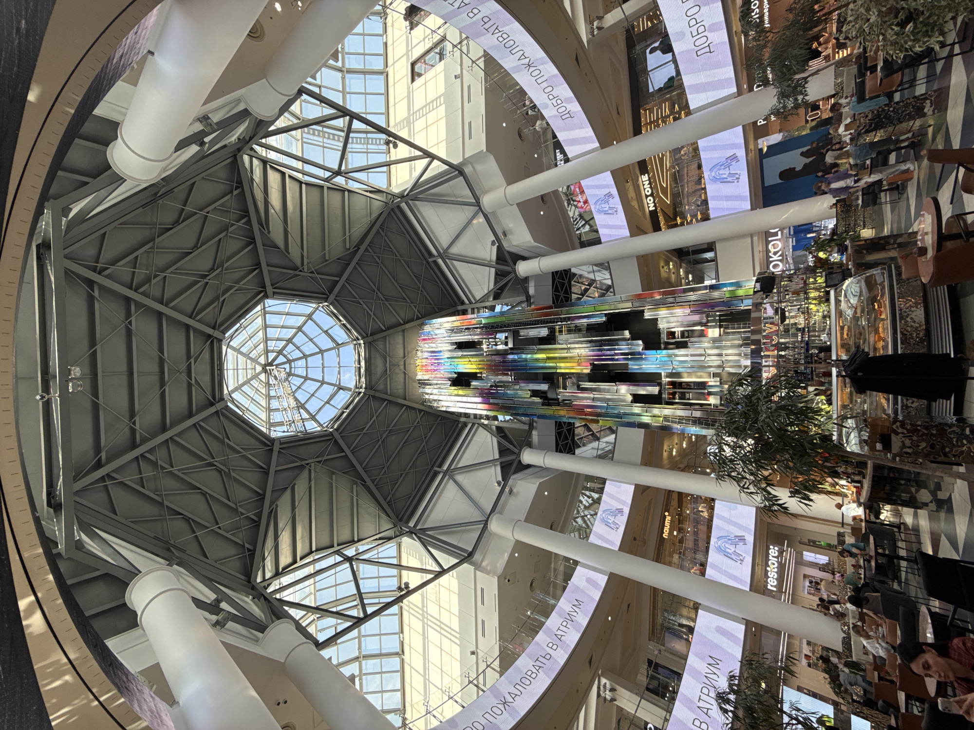 The image shows the interior of a large shopping mall, likely located in Moscow, Russia, based on the Cyrillic text present. The perspective is looking upwards towards the mall's central atrium, which features a complex geometric glass and metal roof structure, letting in natural light. A colorful, multi-layered art installation hangs from the ceiling of the atrium, adding a vibrant focal point.
The atrium is surrounded by multiple levels of shops and businesses, evident from the signage and advertisements visible on the mall's walls. Large white support columns are spaced throughout the atrium. Seating areas with tables and chairs can be seen, with a few people present, suggesting food or beverage services. The atmosphere appears bright, modern, and inviting.