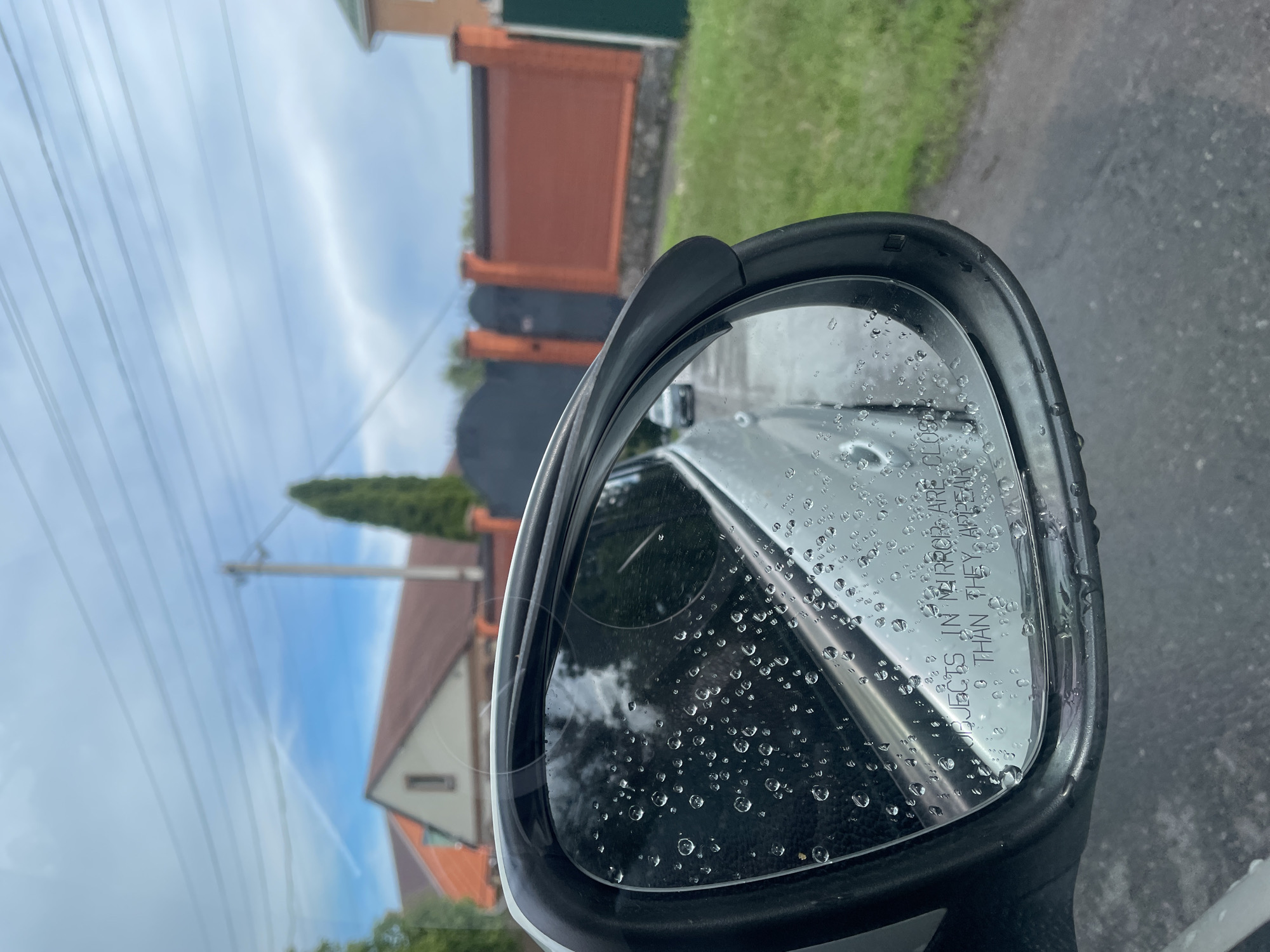 The image shows a side mirror of a car covered in raindrops. The inscription "OBJECTS IN MIRROR ARE CLOSER THAN THEY APPEAR" is visible on the mirror's surface. The reflection in the mirror includes a blurred view of a residential area with buildings, a green lawn, and power lines. The location is possibly in Poltava, Ukraine. The weather appears to be rainy. The road is wet. There are no visible people.