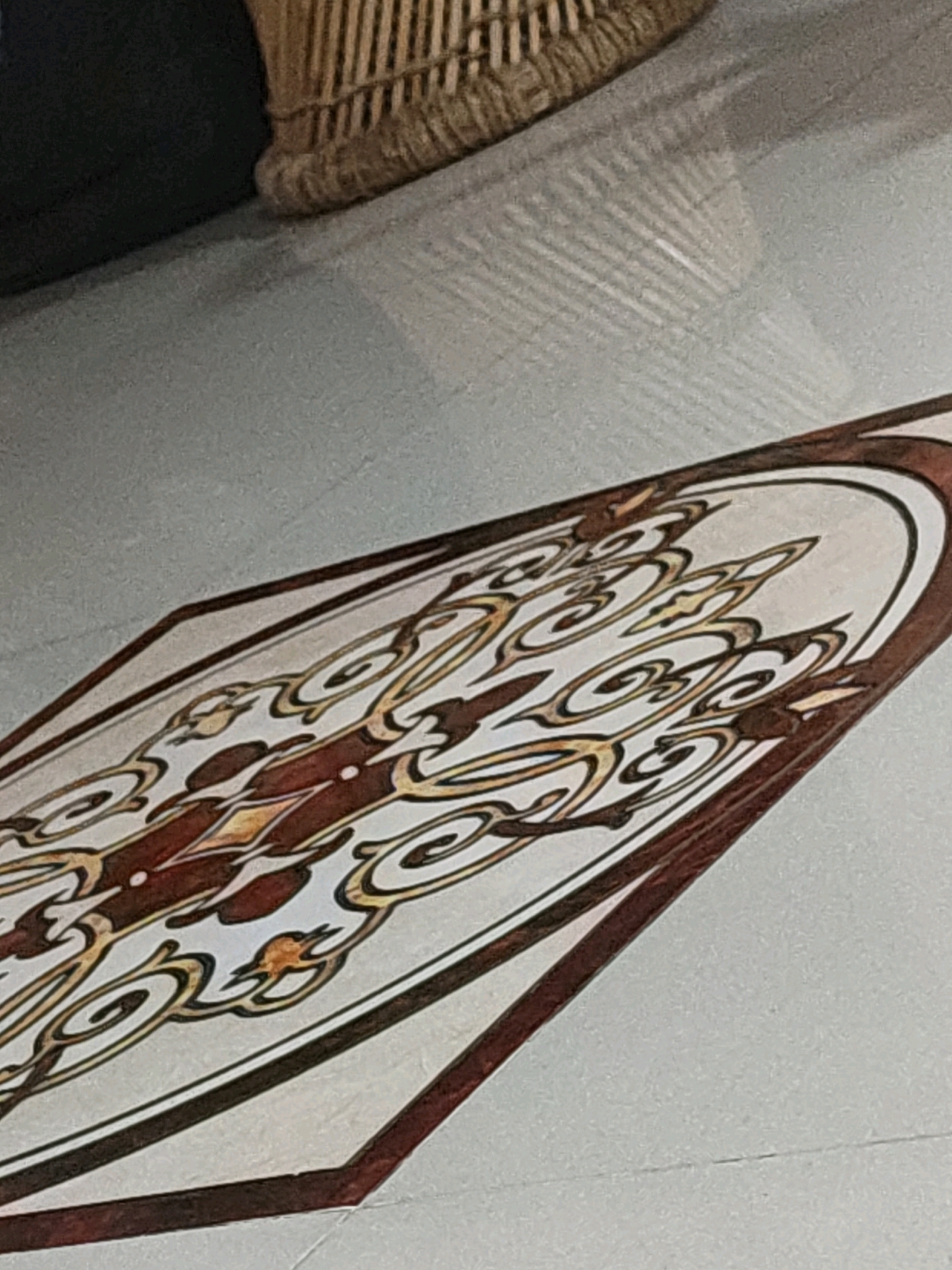 The image shows a section of an ornate tile with a repeating pattern of swirling lines and what appear to be stylized floral or paisley motifs in shades of brown, yellow, and white. The tile is laid on a light-colored floor. In the upper left of the image, a dark object, possibly furniture, and the woven texture of a basket-like object are visible. The floor exhibits a subtle sheen and reflects light, creating linear patterns. The scene appears to be indoors. No specific location in Pune, India, can be identified from the image alone.