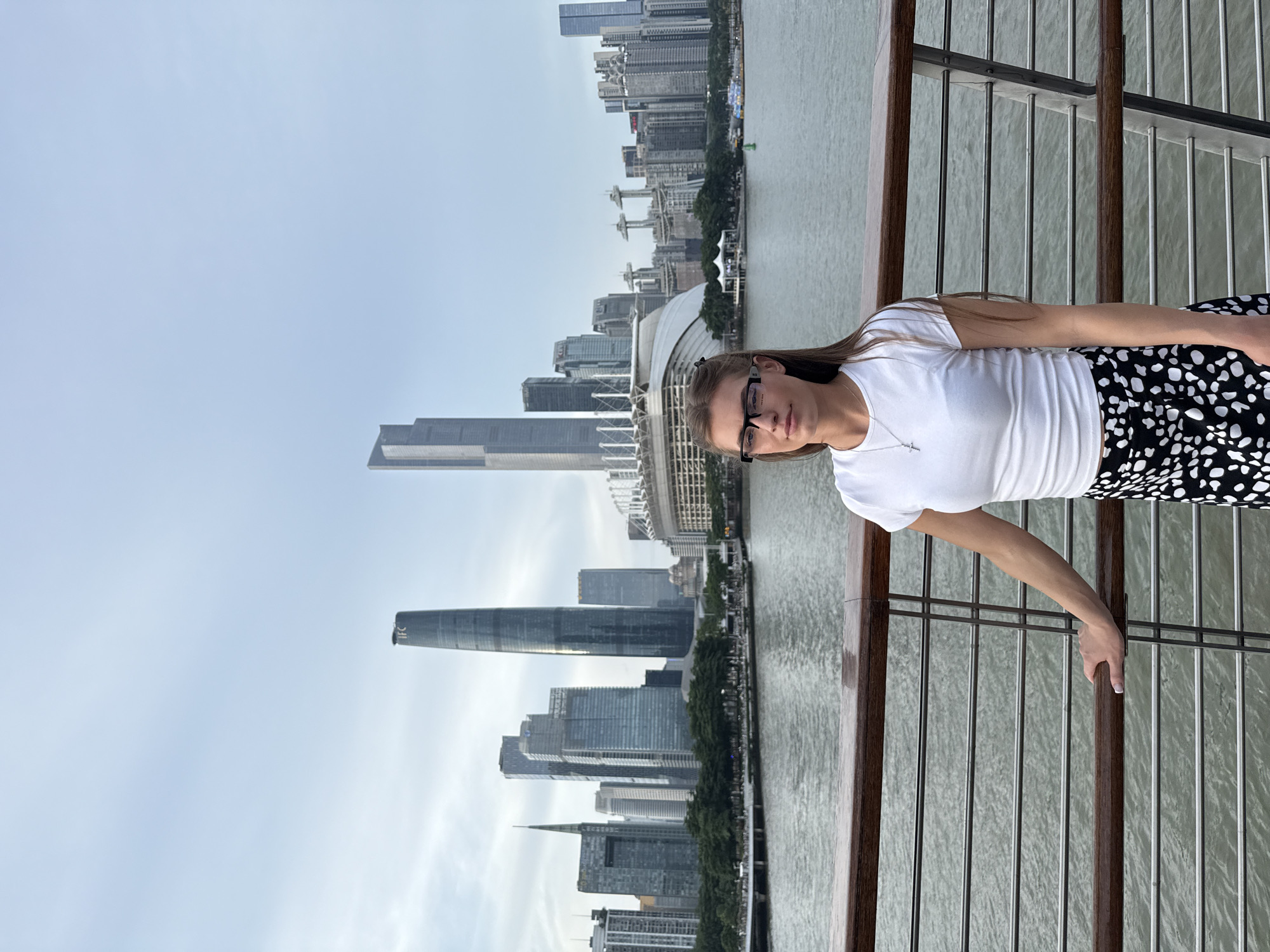 A young woman stands on a railing overlooking a city skyline and a body of water. She is wearing a white t-shirt, black and white polka dot skirt, and glasses. Her left arm is resting on the railing, and her right hand is extended. The background features numerous skyscrapers of varying heights and architectural styles, some appearing to be under construction. A large domed stadium structure is also visible. The sky is overcast, suggesting it might be dawn, dusk, or a cloudy day. The water appears calm, reflecting the sky and buildings. The overall scene depicts an urban environment with a modern aesthetic. The image is oriented vertically, with the woman on the right side of the frame and the cityscape on the left. There is no visible text in the image. The location is identified as Haizhu District, China.