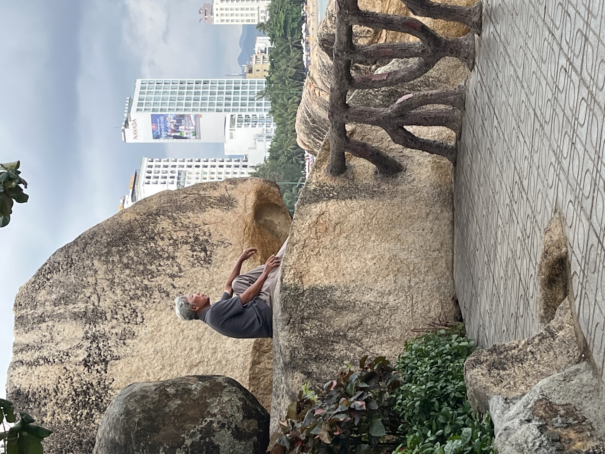 A middle-aged man with gray hair is lying on his back between two large, light-colored rocks. He is wearing a dark gray shirt and light brown pants. In the background, there are tall buildings and palm trees. The location is likely Bac Nha Trang Ward, Vietnam, given the urban and tropical environment. A decorative concrete fence with faux tree branches is to the right. Paved walkway. Plants at base of rocks.
