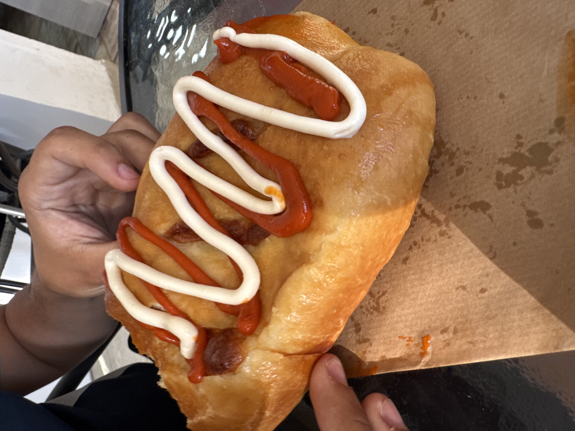A close-up, slightly angled shot depicts a hand holding a pastry covered with white and red sauce. The pastry is golden brown in color and rests on a brown paper surface. Parts of another hand and a chair can be seen in the periphery. The setting appears to be outdoors, possibly a cafe or eatery, due to the visible table surface. The location and city are unknown.