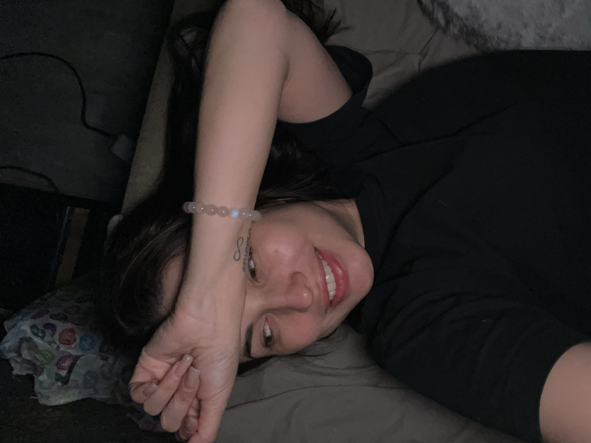 A woman lies on a bed in an indoor environment, wearing a black top. Her left arm is raised with her hand near her forehead. A bracelet adorns her left wrist, and a tattoo is visible on her forearm. To her left is a pillow with a patterned pillowcase. A cable is visible in the background to the left.