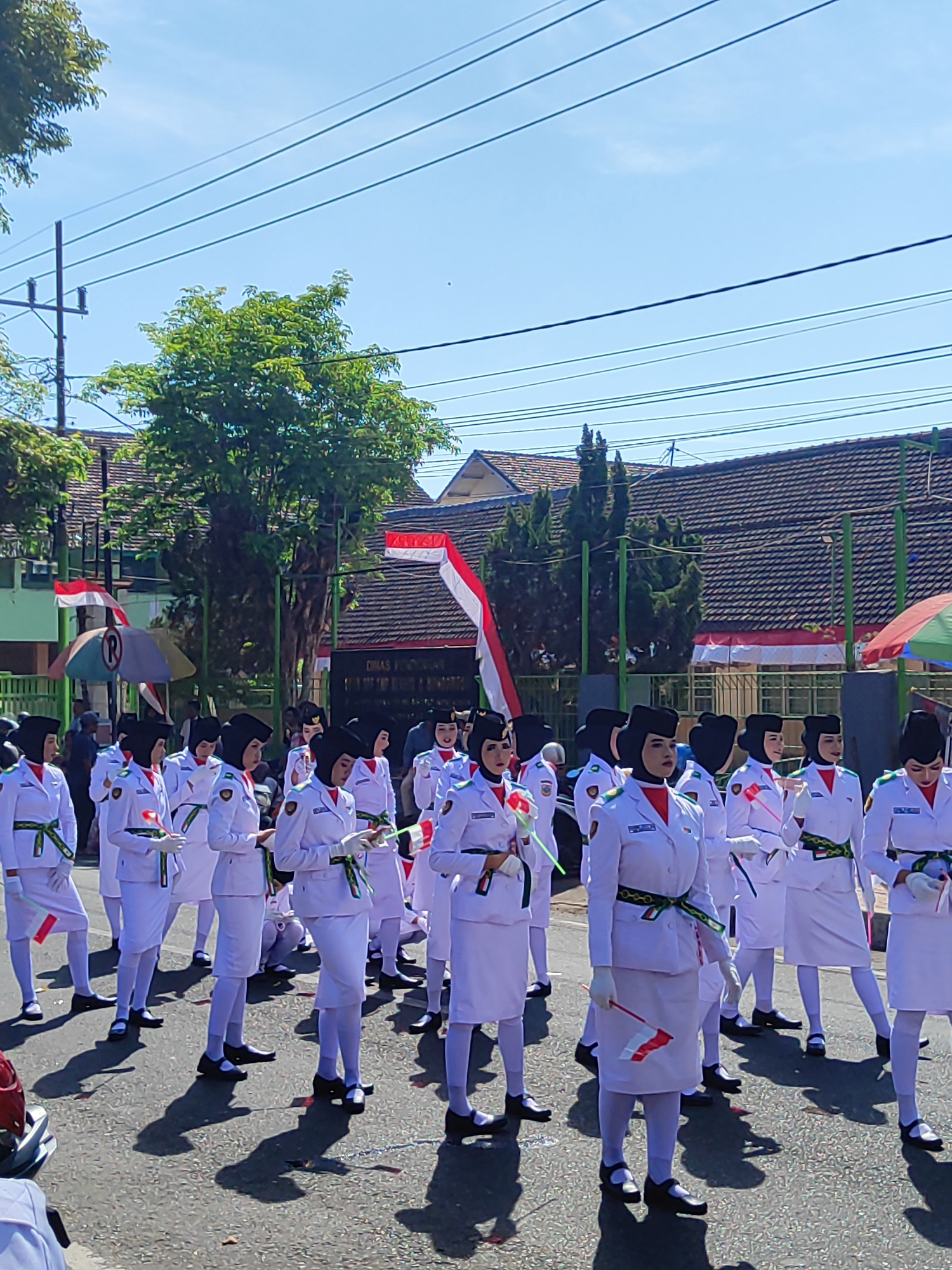 In Dabasah, Indonesia, a group of young women in white uniforms with black head coverings and hats are marching in formation outdoors on a sunny day. The uniforms are accented with red and green trim and sashes. Many of the participants are holding small Indonesian flags. In the background, there are trees, buildings with red tile roofs, and overhead power lines. A building in the background has a sign that reads "DINAS PENDIDIKAN." The scene appears to be a parade or ceremonial event.