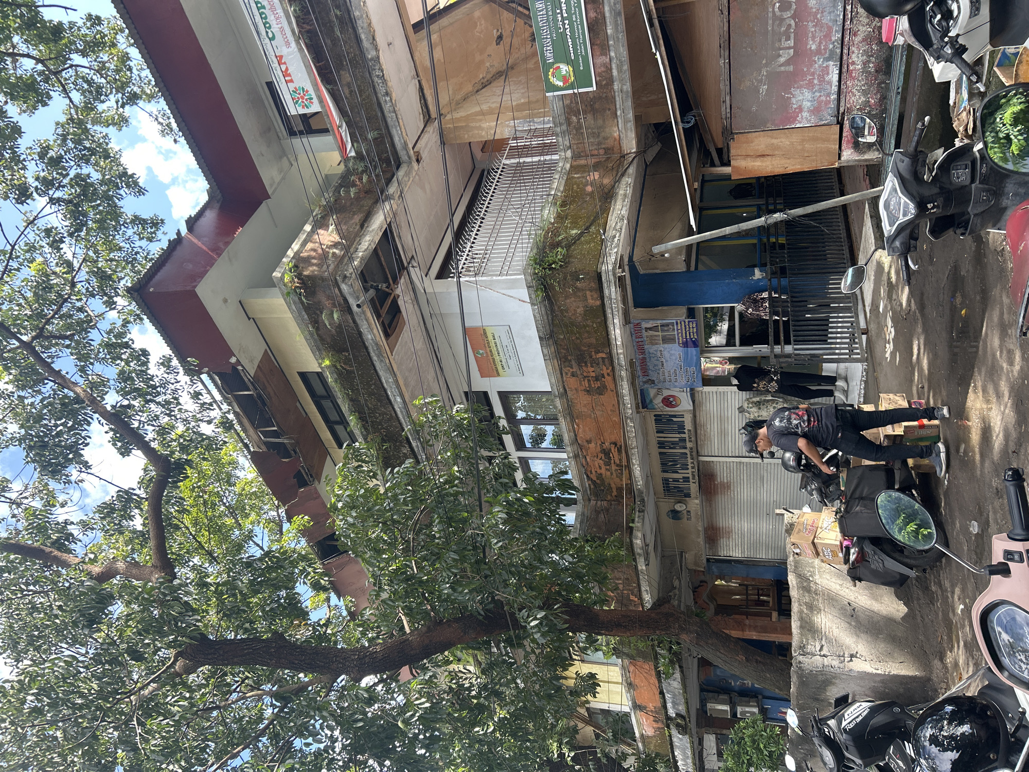 The image shows a street scene in Denpasar, Indonesia. In the foreground, several motorcycles are parked on a dirty street. A man in a black t-shirt and black jeans is standing next to a motorcycle laden with boxes. Behind the man is a multi-story building with several businesses on the ground floor. One of the businesses has a sign that reads "Warteg Puspita Bali Dwipa". The building appears to be old and somewhat neglected, with moss growing on some of the ledges. Overhead, electrical wires crisscross the scene, and a large tree with green leaves partially obscures the view of the sky. The overall impression is one of a busy, everyday scene in an urban Indonesian environment.
