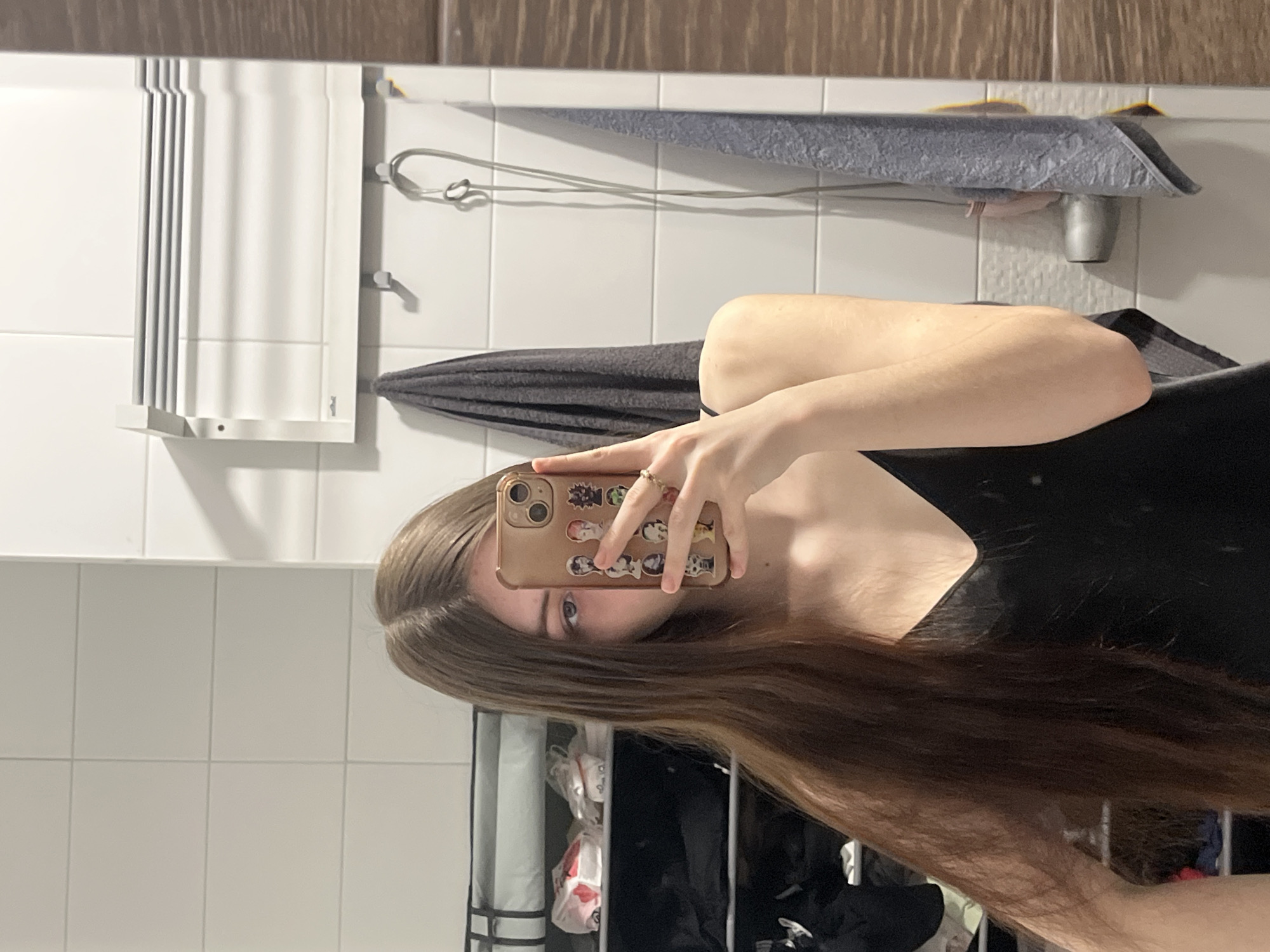 A selfie, taken indoors, with the subject positioned in a bathroom. The subject is holding a mobile phone, decorated with stickers, partially obscuring their face. The subject has long, brown hair and is wearing a black top.
The bathroom features white tile walls, a towel rack, a folded gray towel, a shower head and a gray towel hanging above it, and a white, accordion-style towel warmer. The location is not Saint Petersburg, Russia.