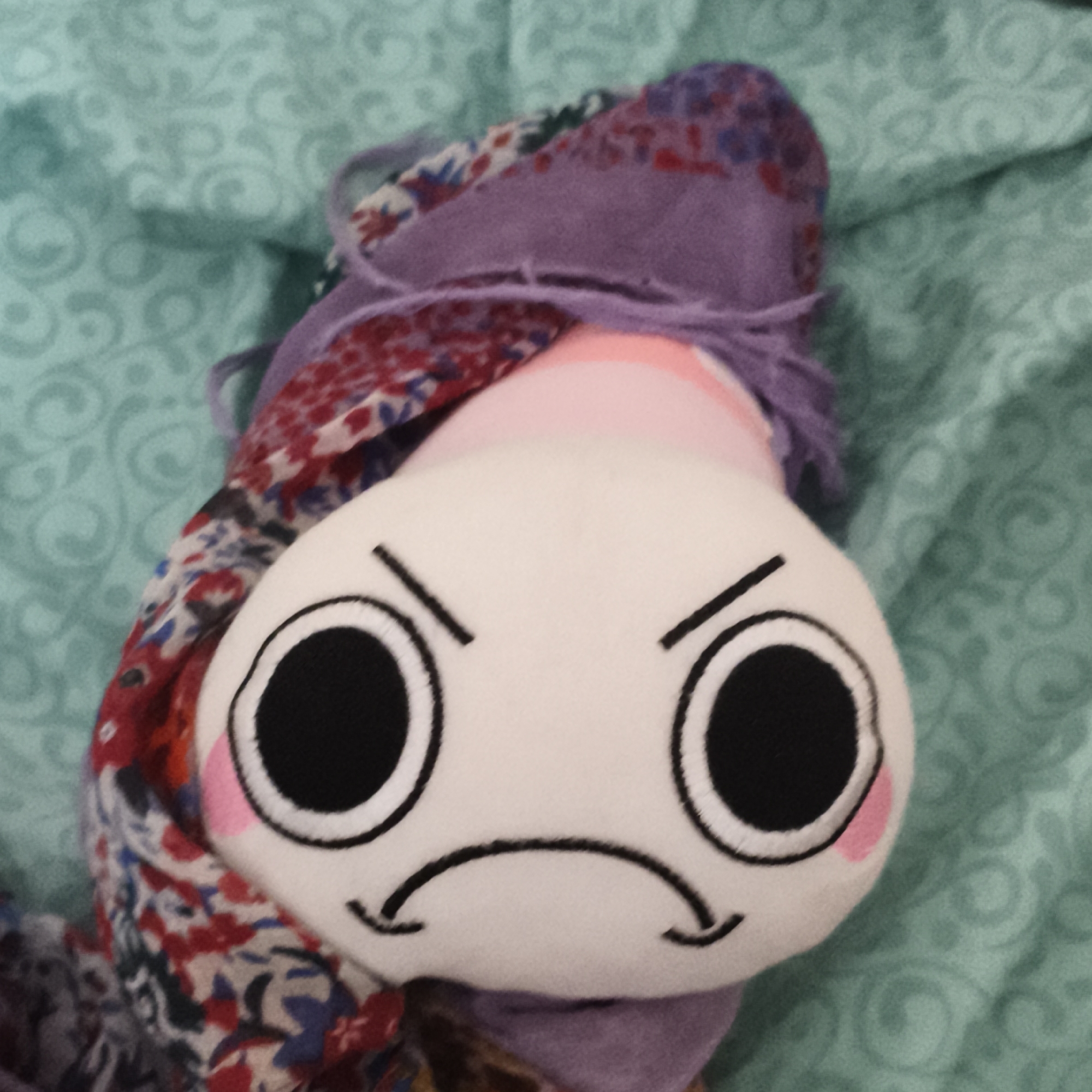 A close-up shot features a plush toy with an angry expression, comprised of a white, round head with large, black, circular eyes outlined in white, pink cheek blushes, black angular eyebrows, and a downturned black mouth. The plush is wrapped in a colorful scarf with floral patterns and a purple wool scarf, both of which are draped around the plush's head and neck. The background consists of a patterned, aqua-colored fabric, which appears to be a blanket or sheet.