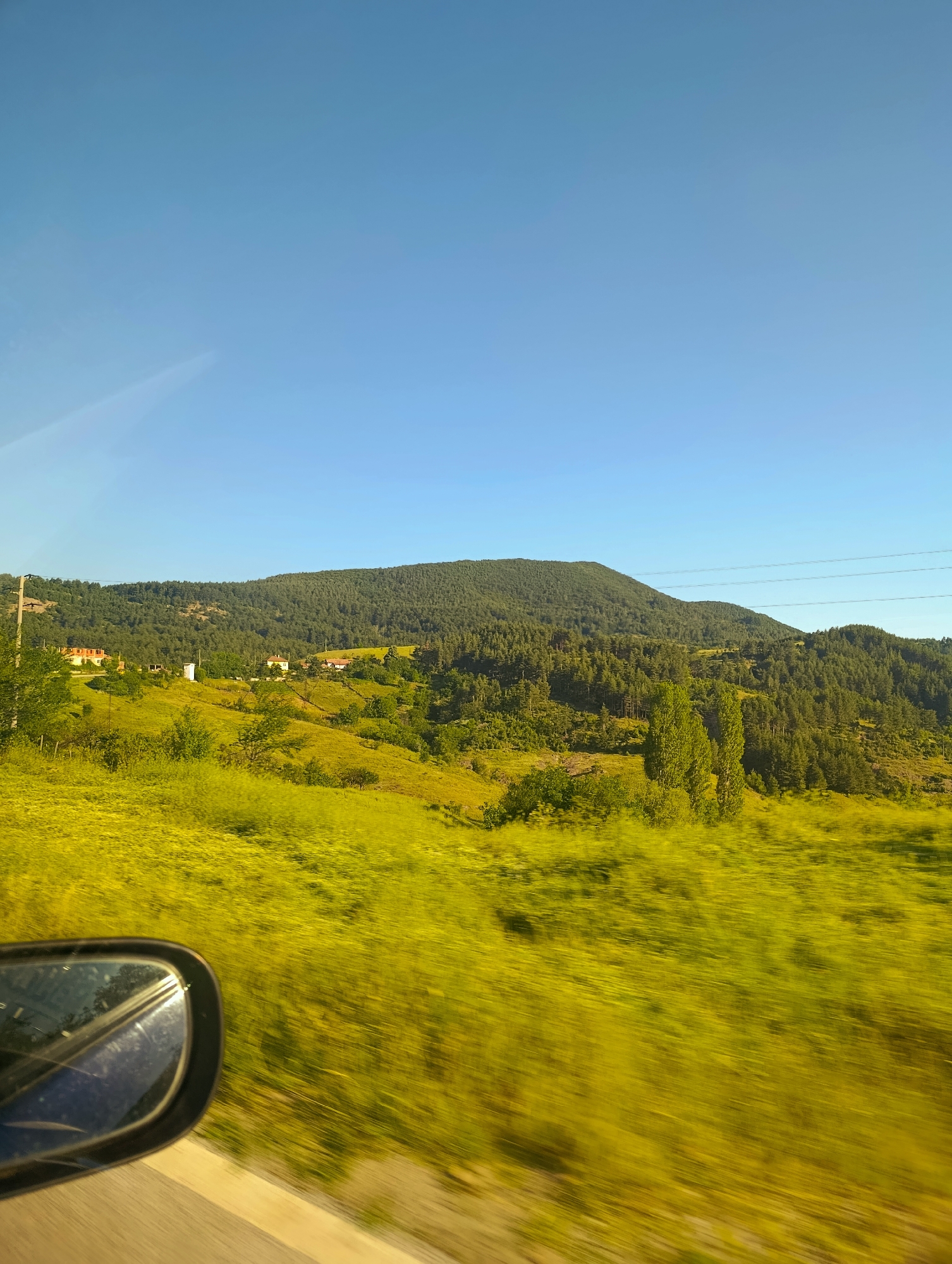 The image captures a scenic view of rolling hills covered in lush green vegetation, with a dense forest dominating the background. The sky is a clear, vibrant blue. In the mid-ground, several houses are nestled among the greenery, suggesting a rural or semi-rural settlement. The foreground is a blur of bright yellow, indicating fast movement, likely from a vehicle. A side-view mirror of a car is visible on the left, reflecting the surrounding environment. The scene is bathed in sunlight, casting shadows and highlighting the textures of the landscape. This view is characteristic of the countryside surrounding Komuniga, Bulgaria, known for its rolling hills and forested slopes.