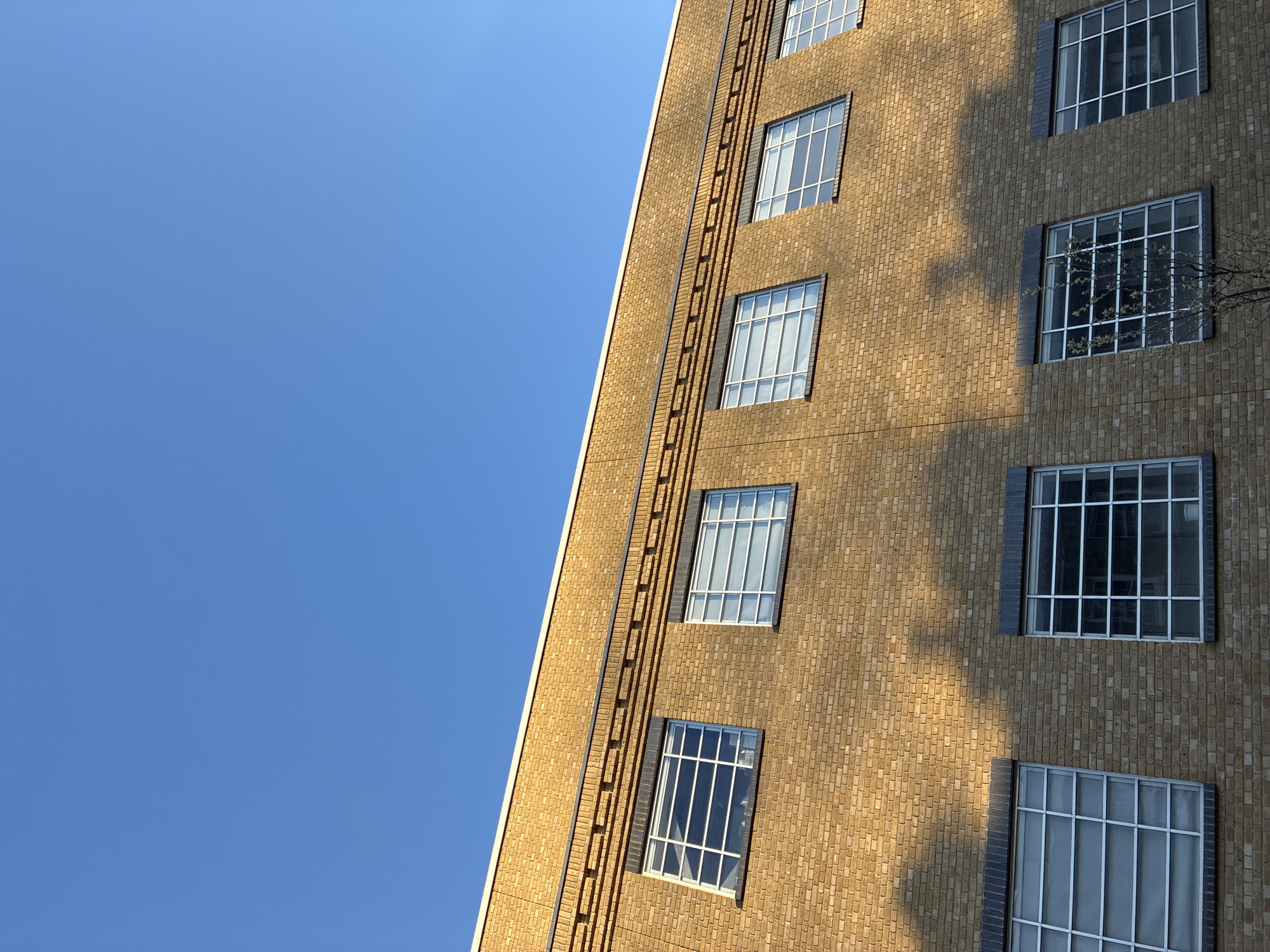 A low-angle shot of a building in London, United Kingdom shows a tan-brick facade with evenly spaced windows and a fire escape. The building extends upward from the lower-right corner of the frame, leaving a large expanse of clear, blue sky in the upper-left. The windows have multiple panes and are framed in a dark gray material. The building is illuminated by the sun, which casts shadows on the brickwork. A fire escape ladder is visible running vertically along the building's edge. There are no people visible in the image.