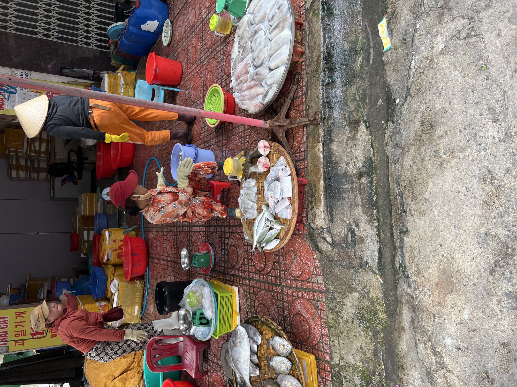 The image captures an outdoor market scene in Nha Trang, Vietnam, likely near the coast given the prevalence of fresh seafood. Two women are present. One woman, wearing a red cap, orange and white patterned shirt, and white gloves, is seated on a red stool, attending to a display of fish and squid. She is handling what appears to be prepared fish fillets. In front of her is a large round tray laden with various types of squid. To her left, a woven tray holds several whole fish. Another woman, wearing a brown cap, orange pants, and yellow gloves, stands facing away from the camera, wearing a conical hat. She is positioned behind stacked boxes and crates, some of which are yellow and wrapped in plastic. The ground is paved with red and brown patterned tiles. Several plastic buckets, tubs, and scales are visible, along with a bright yellow sign in Vietnamese. The overall impression is a bustling, working market environment with a focus on the sale of fresh seafood.