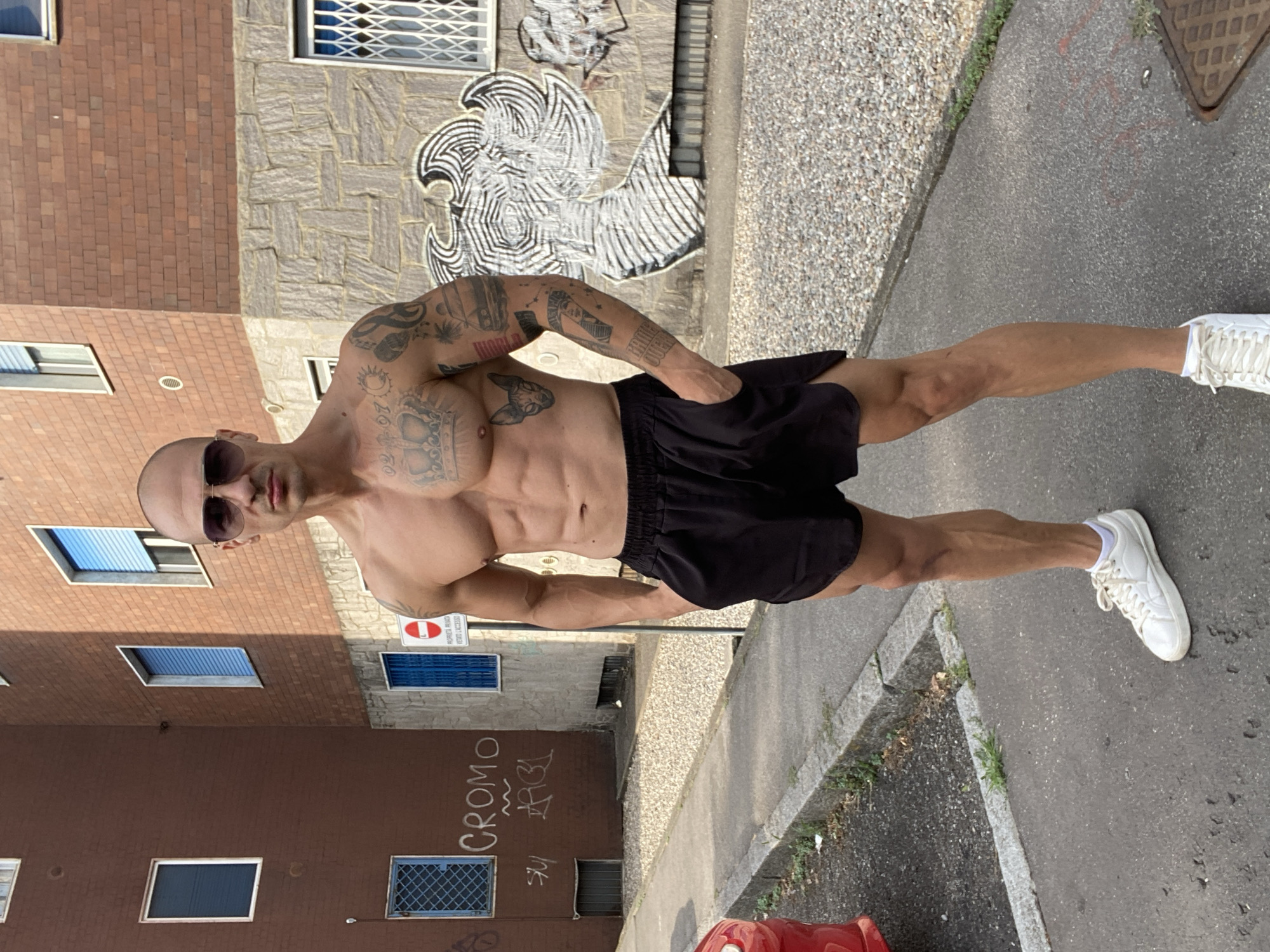 The image shows a muscular, bald man with sunglasses and tattoos on his torso and arms, wearing black athletic shorts and white sneakers. He is standing on a paved sidewalk in front of a brick building with graffiti on its lower section. The building has several windows, some with blue shutters. A red car is partially visible on the left side of the image. The man's hands are in his pockets. The scene appears to be in an urban or suburban environment. The graffiti on the building includes the word "CROMO". The man's posture and attire suggest he may be engaged in or have just finished physical activity.