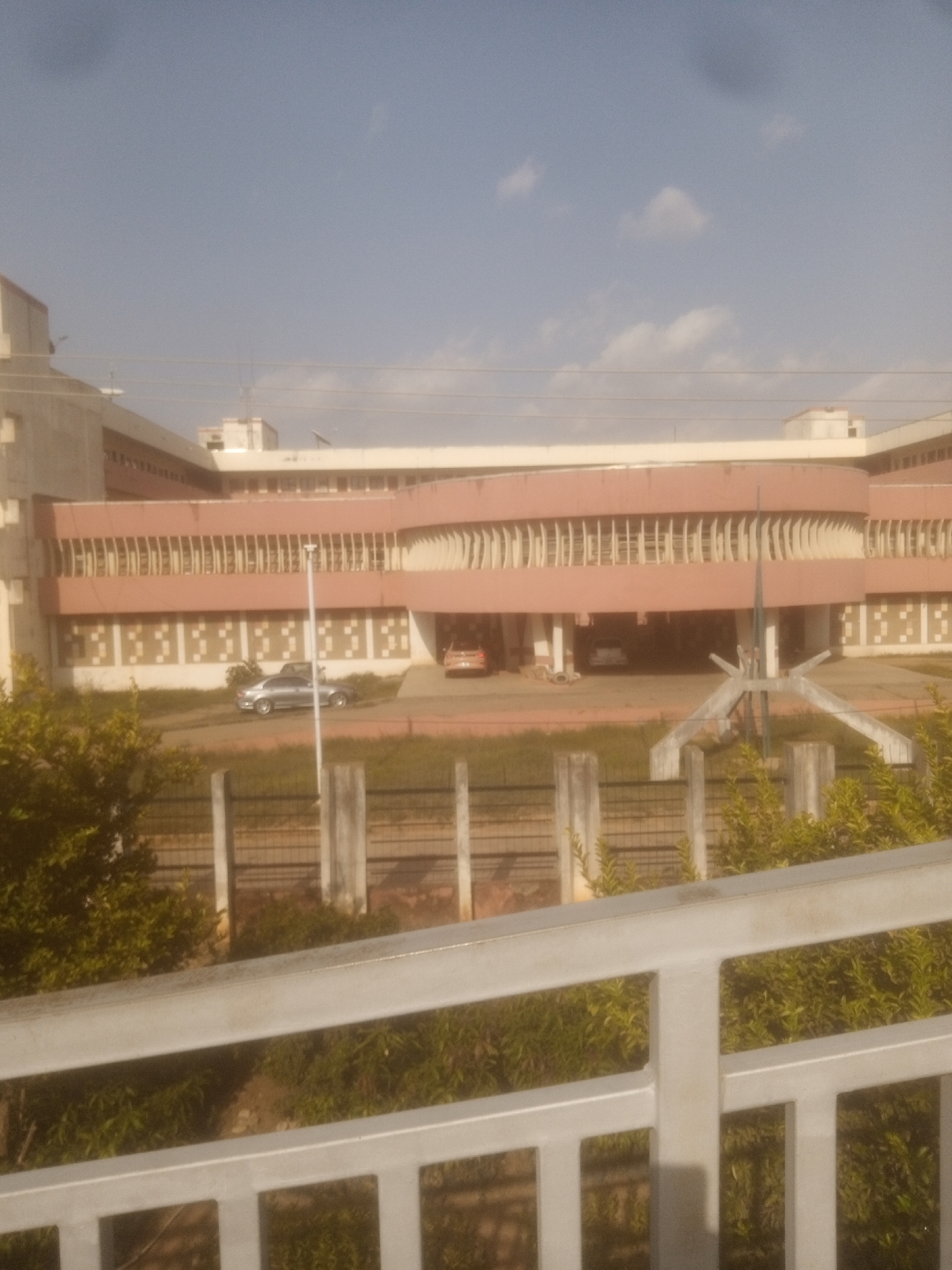 The image depicts a large, modern building with a distinctive pink facade and numerous vertical window elements. The building appears to be a significant public or institutional structure. In front of the building, a silver sedan is parked, and further to the right, a portion of another vehicle is visible. The foreground is partially obscured by a railing, and behind it, lush green foliage and some fence posts are present. The sky above is a pale blue with scattered clouds. The scene suggests an outdoor setting in a developed area, likely during daylight hours. The image is likely of a location in Unguwan Dosa, Nigeria, given the context provided.