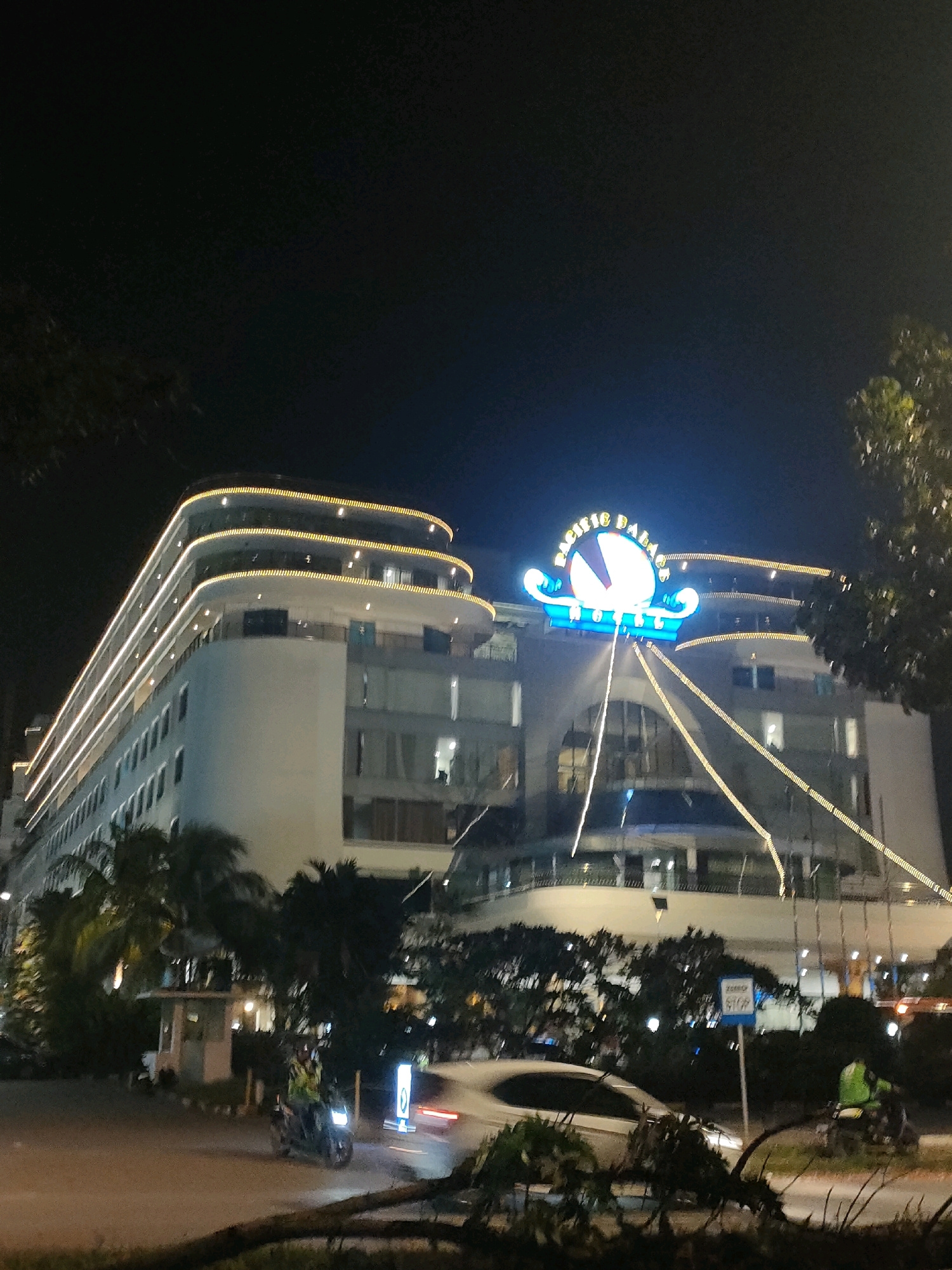 The image depicts the Pacific Palace Hotel in Batam, Indonesia, at night. The hotel is a multi-story building with a curved facade, illuminated by strings of golden lights along its balconies and edges. A prominent neon sign displaying "PACIFIC PALACE HOTEL" in blue and yellow is visible at the top of the building. The scene includes several palm trees and other foliage in the foreground and around the hotel. On the street in front of the hotel, a person is riding a scooter, and a white car is parked. Streetlights and building lights contribute to the nighttime ambiance. The sky is dark, indicating it is nighttime.