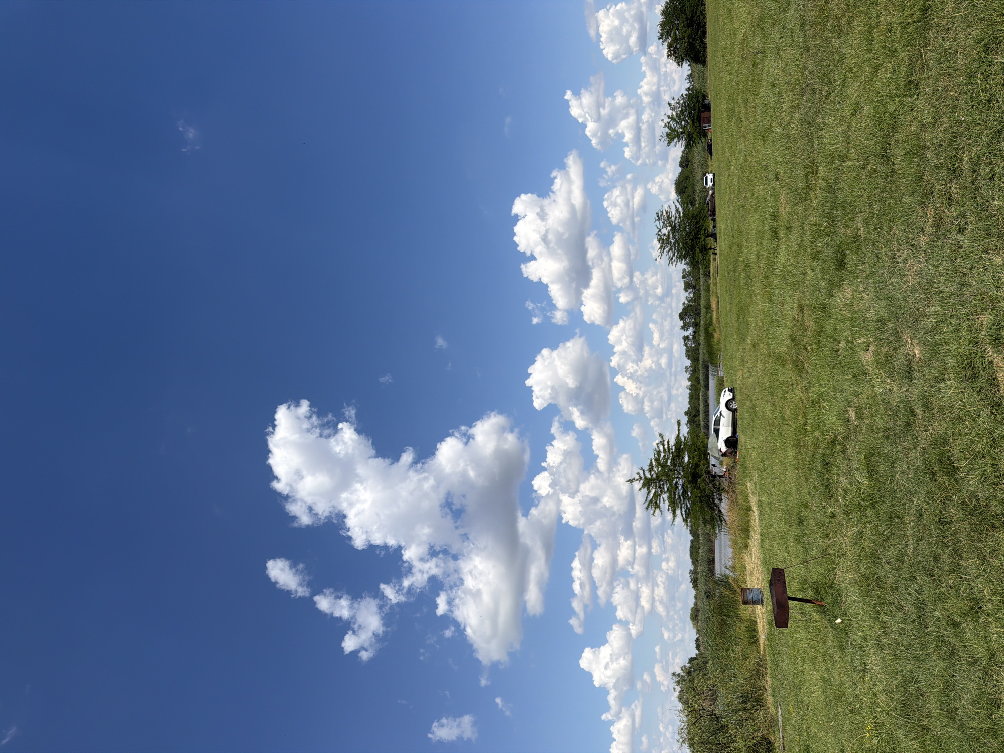 This is a horizontal, eye-level, outdoor shot of a grassy field with a bright blue sky. The sky is filled with fluffy white clouds. In the background, there is a body of water with trees and vegetation on the far bank. A white car is parked on the grassy area near the water. There is also a metal barbecue grill in the foreground. The sun is shining, indicating it is daytime. The scene appears to be a park or a recreational area. There is no visible text.