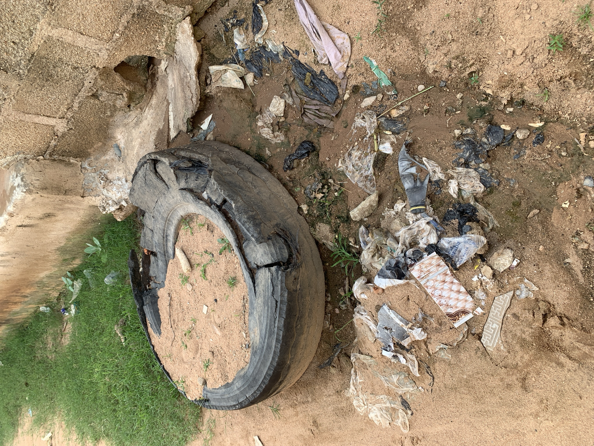 In an outdoor, urban environment, a worn-out tire filled with sand lies on the ground. Scattered around the tire is a significant amount of litter, including various plastic bags, wrappers, and what appears to be a broken piece of ceramic tile. The ground is a mix of sandy soil and dirt, with some sparse green vegetation growing in patches. In the background, a rough-textured wall made of concrete blocks stands, with some areas showing signs of weathering and damage. A patch of overgrown green grass is visible to the left of the scene, adjacent to the wall. There are no people or animals present in the image.