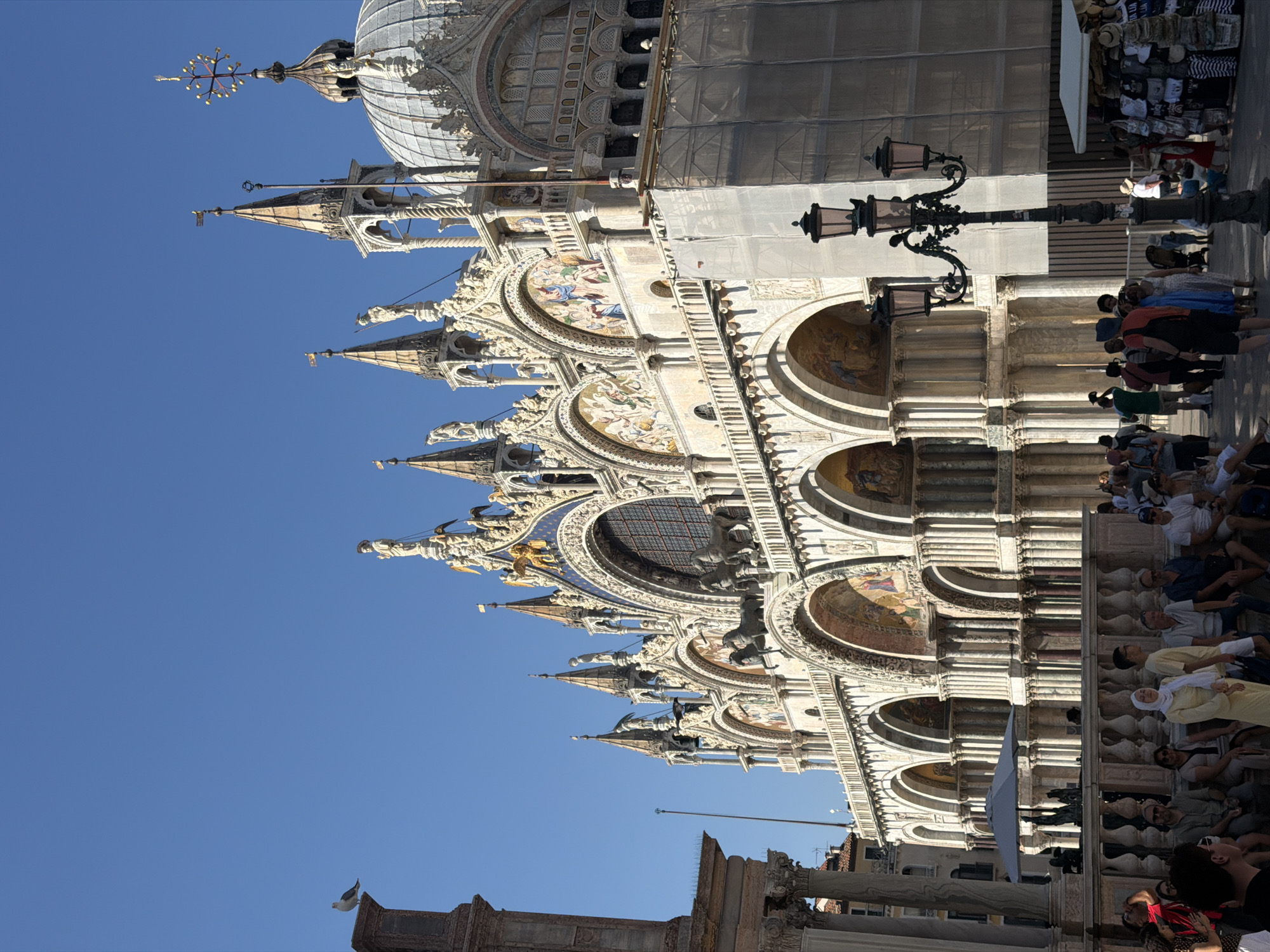 The image depicts a sun-drenched scene in St. Mark's Square in Venice, Italy. The magnificent Basilica di San Marco, a UNESCO World Heritage site, dominates the frame with its ornate facade featuring multiple domes, spires, intricate carvings, and mosaic-adorned arches. The basilica's upper levels are partially covered with scaffolding, indicating ongoing restoration work. In the foreground, a lamppost with three ornate lamps stands casting shadows. A crowd of tourists and locals are visible in the square, some seated on a low wall, others standing and observing the architecture or browsing stalls of souvenirs. A gray umbrella shades an area to the left. The sky above is a clear, bright blue, and the overall atmosphere suggests a warm, bustling day in the heart of Venice.