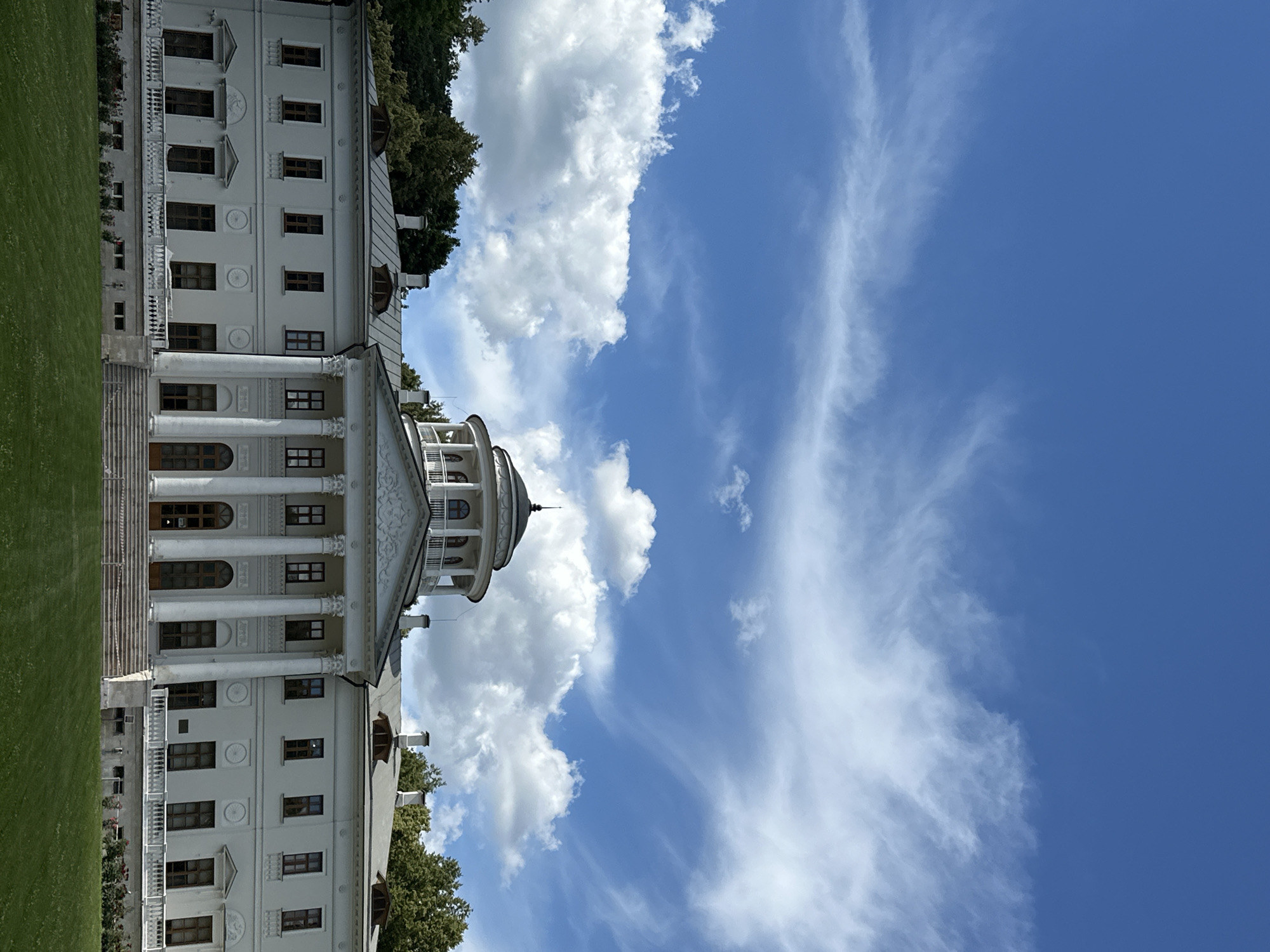 This is a vertical, full-frame, eye-level shot of a grand white classical building, likely a historical estate, with a bright blue sky filled with wispy white clouds. The building features a portico with large white columns, a balustrade, and multiple windows on its façade. A domed structure with arched windows is visible on the right side of the building. The building is partially surrounded by trees and a manicured green lawn. The sun is shining, indicating it is daytime. The weather appears to be pleasant with a mix of sun and clouds. The overall mood is serene and grand, suggesting a visit to a place of historical or cultural significance in Ostafievo, Russia. There is no visible text in the image.